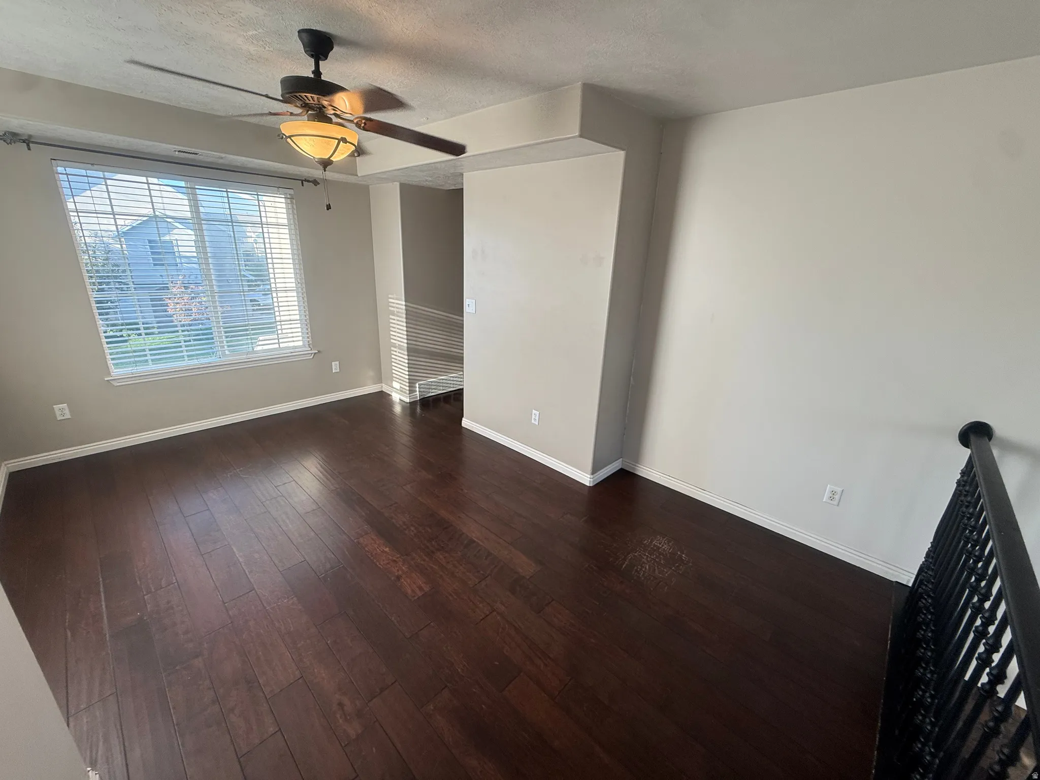 Dark wood-finish flooring, ceiling fan with integrated lighting, large window with blinds, wrought iron railing, and neutral wall paint