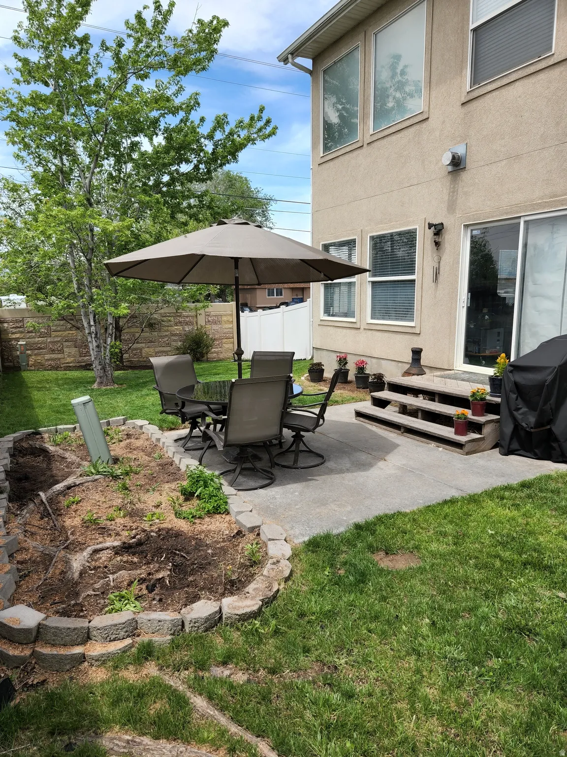 Expansive backyard featuring a concrete patio with steps, a raised garden bed with stone edging, and mature trees. This is a former picture showing what it could look like again...no grass currently.