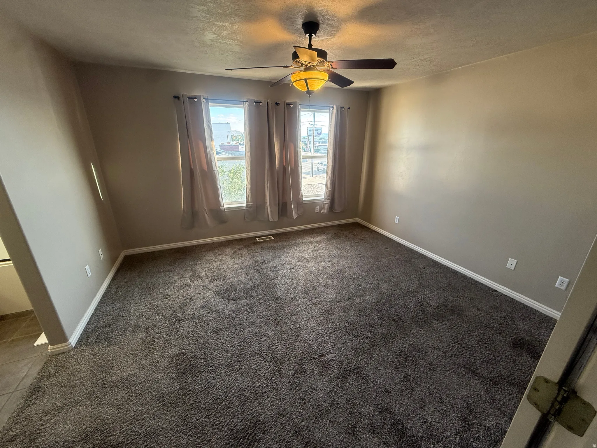 Neutral-toned walls, three windows with drapes, ceiling fan with integrated lighting, textured carpeting, and white trim