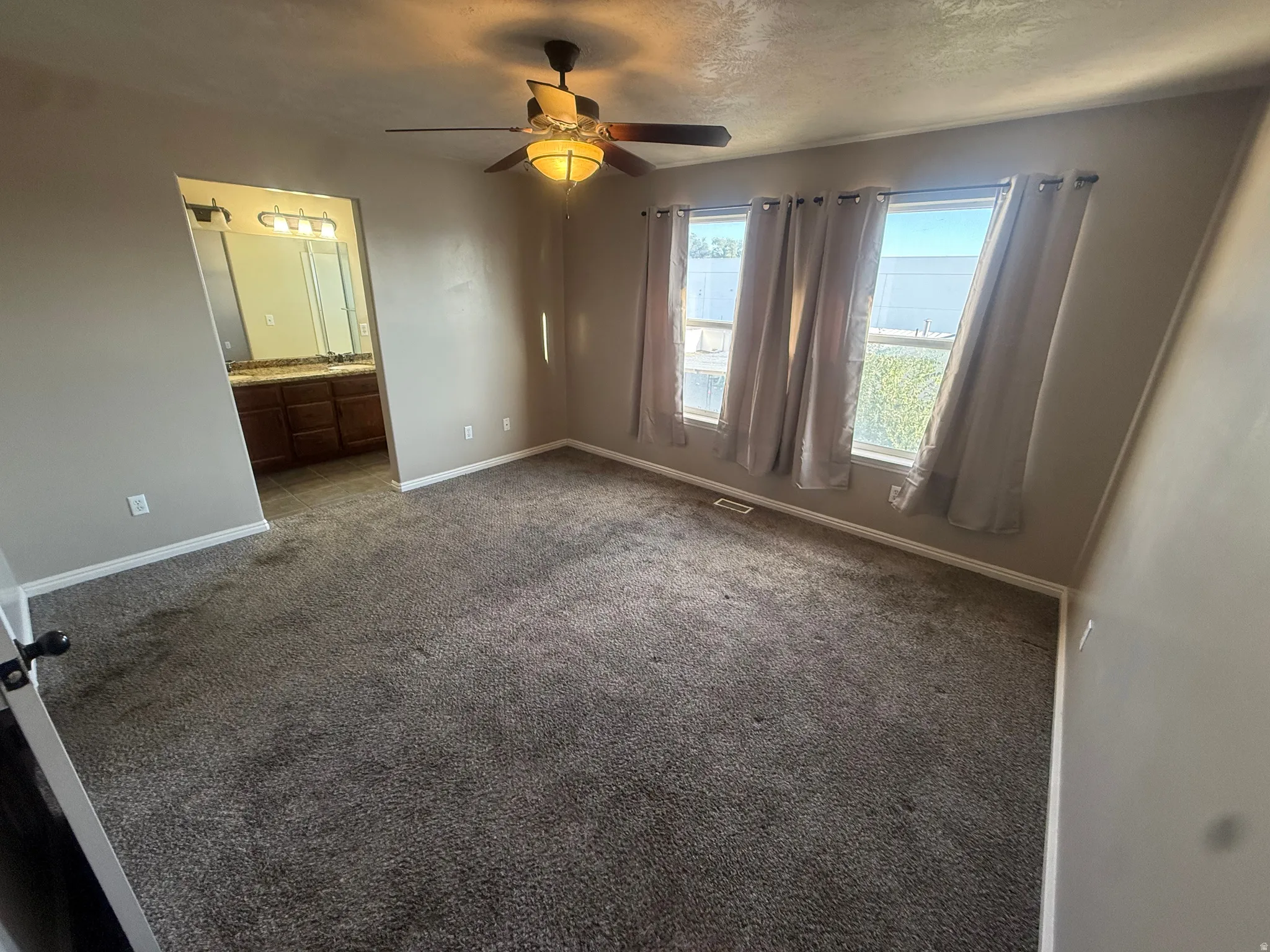Carpeted room with multiple windows, a ceiling fan with integrated lighting, and an adjacent bathroom featuring a dual vanity with a countertop and under-cabinet storage