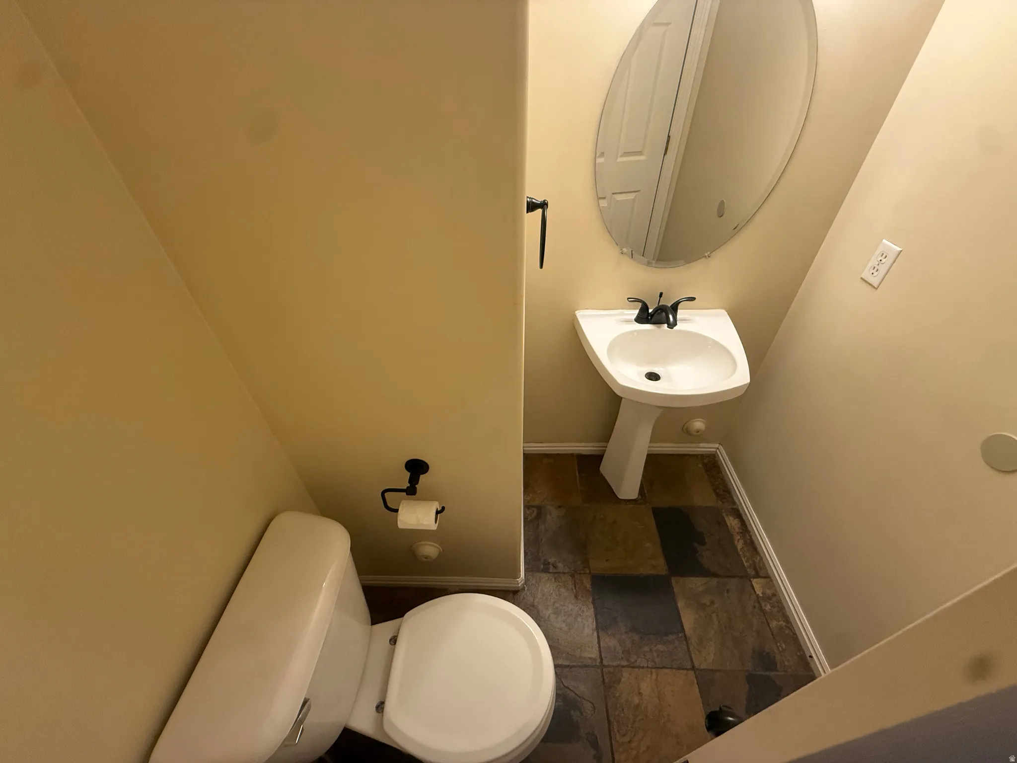 Pedestal sink with dark-toned fixtures, oval wall mirror, and multi-toned tile flooring