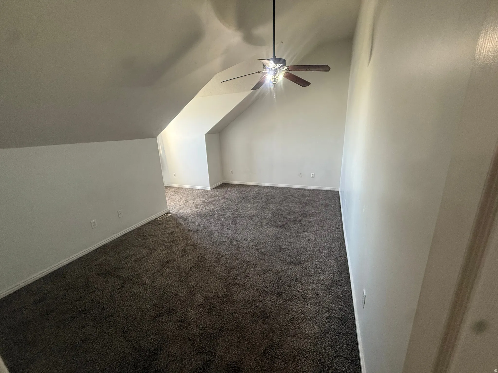 Spacious room with sloped ceilings, plush carpet flooring, a ceiling fan with integrated lighting, and neutral wall finishes