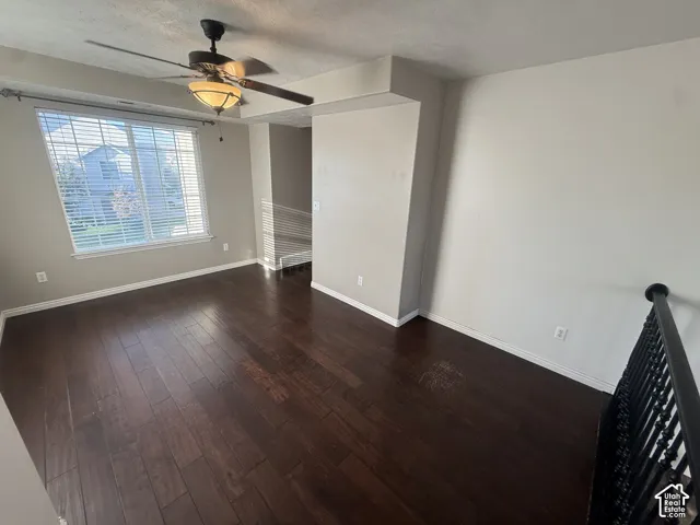 Dark wood-finish flooring, ceiling fan with integrated lighting, large window with blinds, wrought iron railing, and neutral wall paint