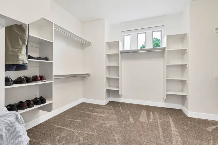 Spacious walk-in closet featuring extensive built-in shelving, multiple hanging rods, durable carpeting, crisp white walls, and natural light from a transom window