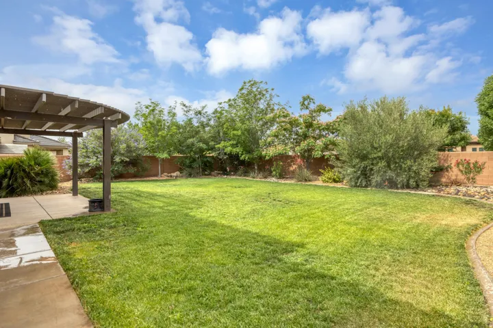 Spacious backyard featuring a curved pergola, expansive lawn, concrete patio, mature trees, and a block wall perimeter