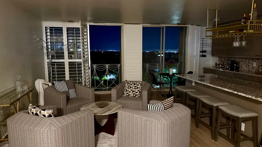 Open living area with wood-finish flooring, a built-in bar featuring a gold-finish hanging wine rack, an extended countertop with seating for four, and exterior access to a balcony through sliding glass doors