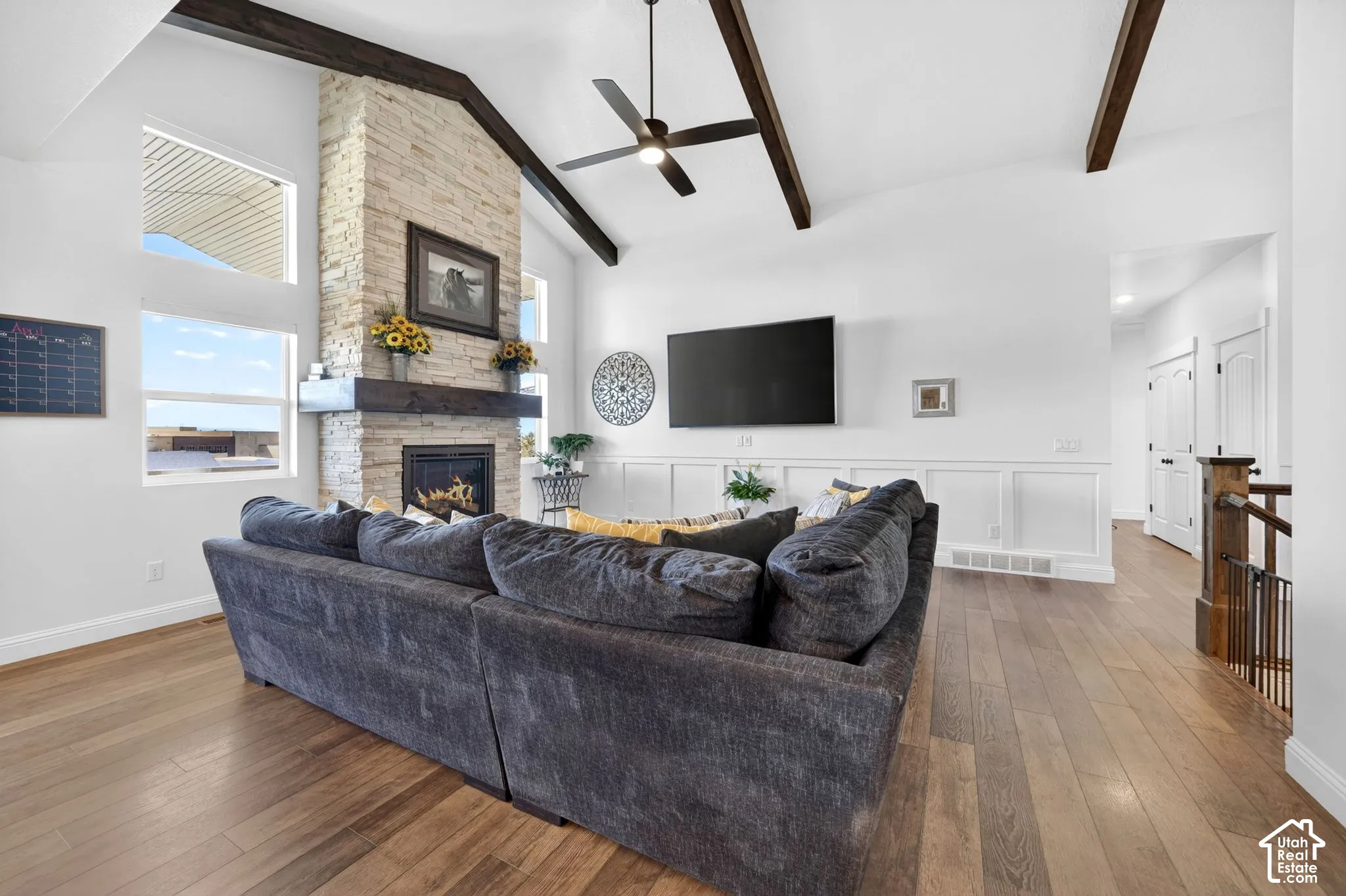 Vaulted ceiling with exposed wood beams, stone-clad fireplace with wood mantle, multiple windows, wood-finish flooring, and wainscoting