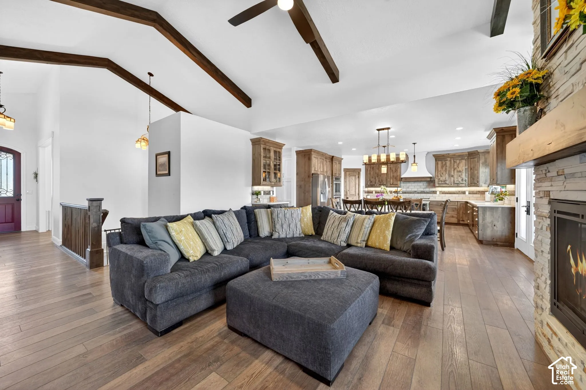 Spacious open-concept living area featuring wood-finish flooring, vaulted ceilings with exposed wood beams, and a stone-clad fireplace with a wood mantle