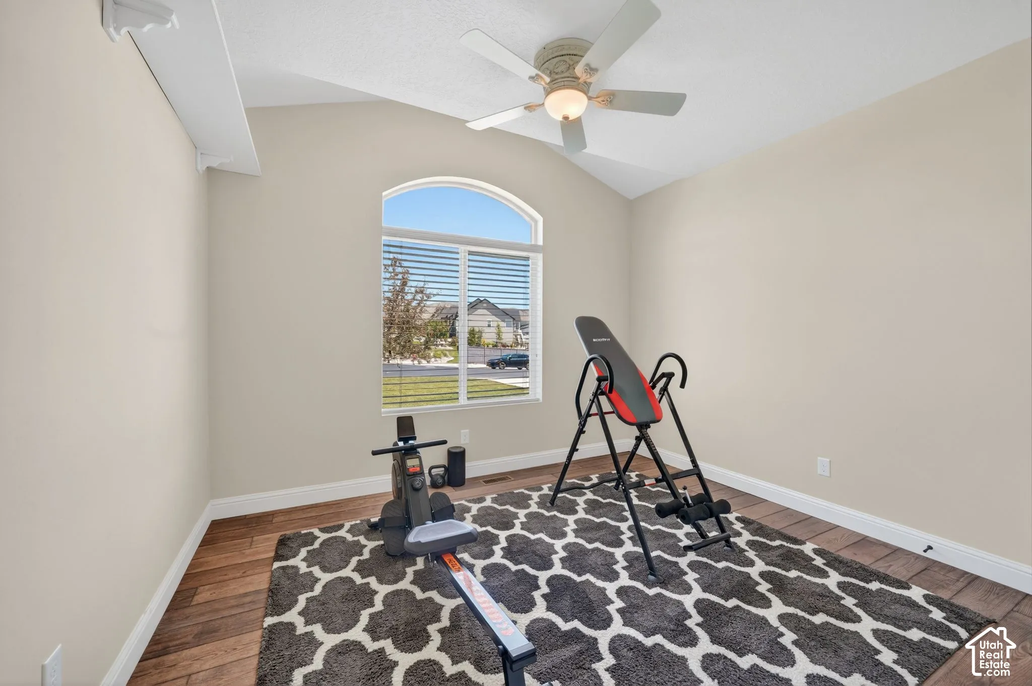 Vaulted ceiling room featuring a large arched window with horizontal blinds, wood-finish flooring, and a ceiling fan with light fixture
