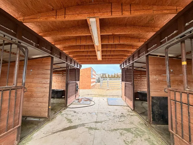 Covered equestrian stalls featuring wood paneling, metal gates, and an exposed wood ceiling with fluorescent lighting