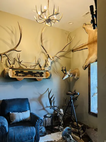 Recessed lighting and antler chandelier illuminate a room featuring wall-mounted antlers, a carved wooden shelf, and a dark leather armchair