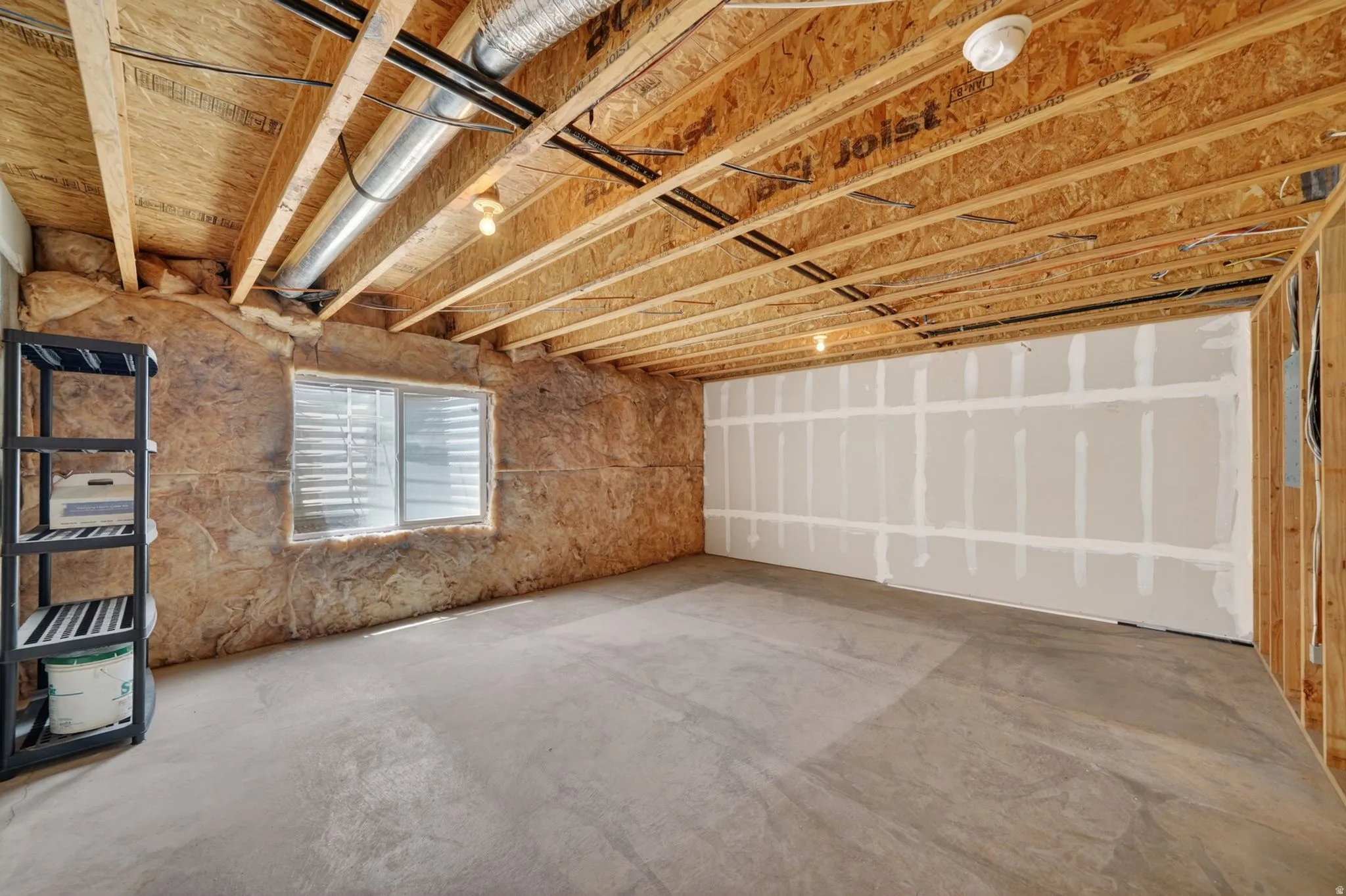 Unfinished basement space featuring exposed wood joists and subfloor, a partial drywall installation, concrete flooring, and a basement window