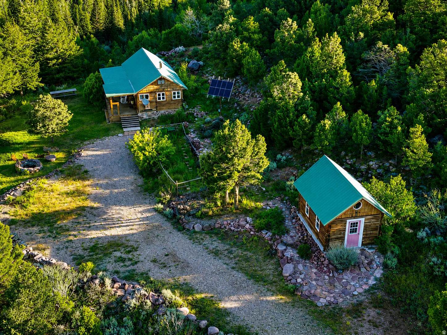 Rural property featuring two wood-sided structures with green metal roofs, a gravel driveway, solar panels, and surrounding mature trees