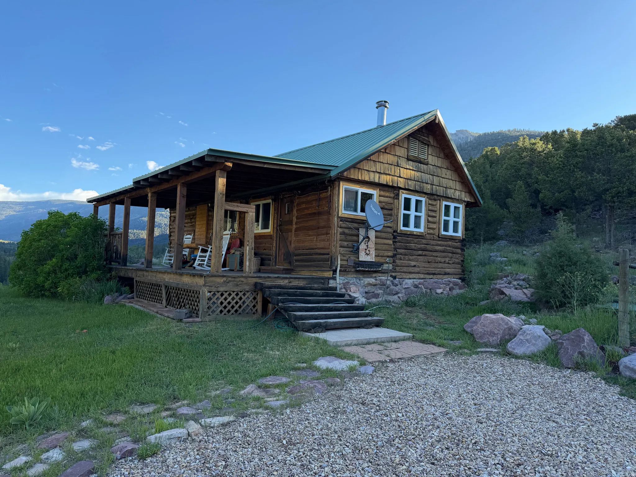 Rustic log cabin exterior featuring a green metal roof, extensive wrap-around porch, and a stone foundation