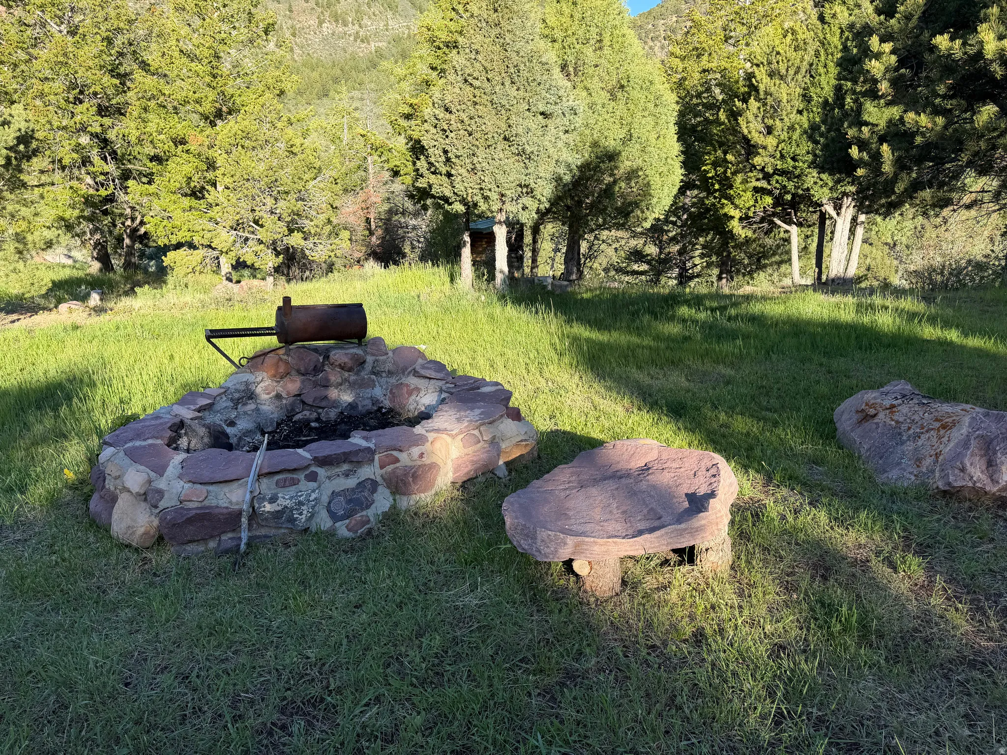 Stone-built fire pit featuring a grilling grate, surrounded by natural rock seating and lush greenery