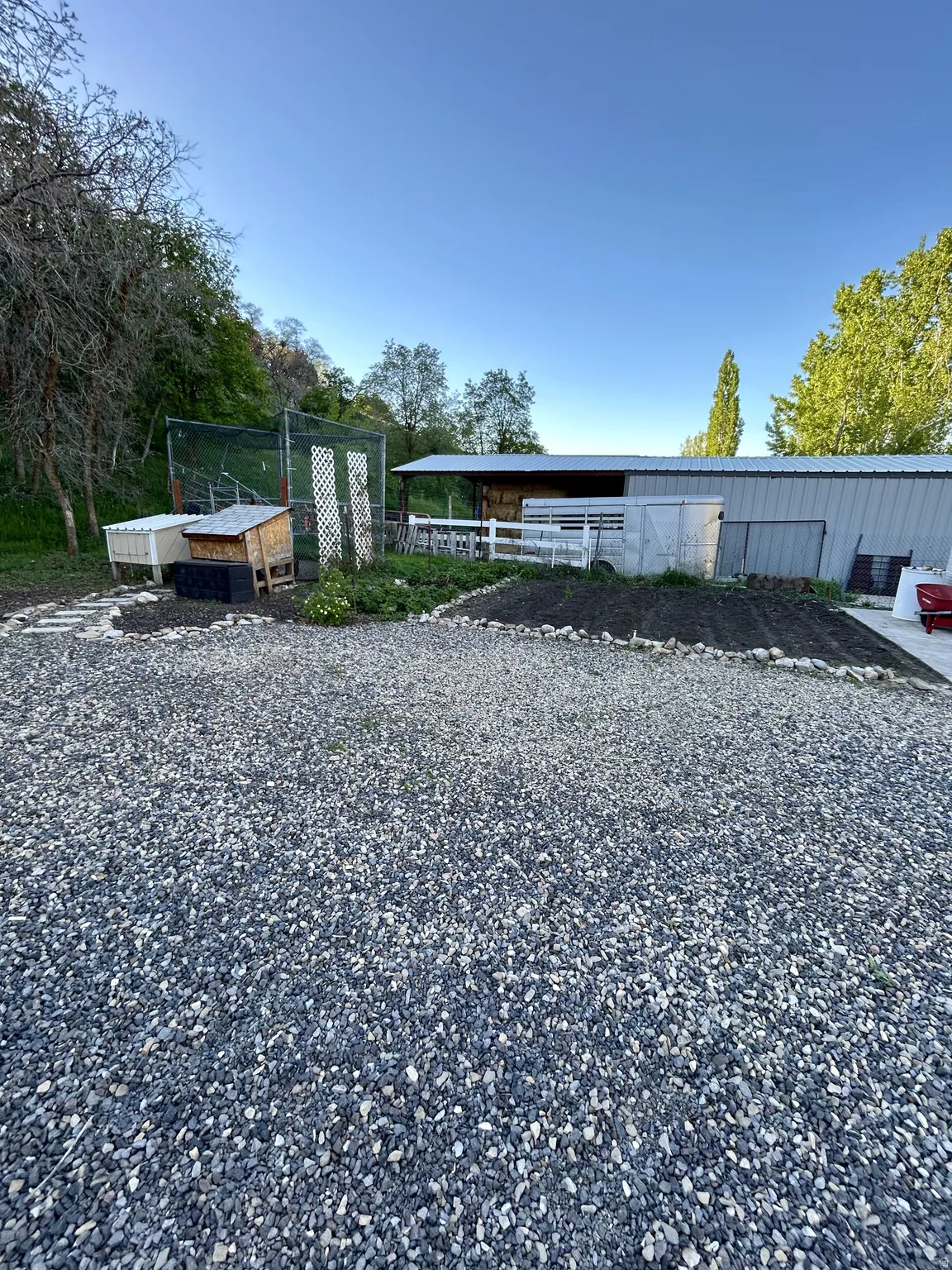 Gravel driveway leading to a metal-sided outbuilding with a lean-to extension, white fencing, and an established garden bed