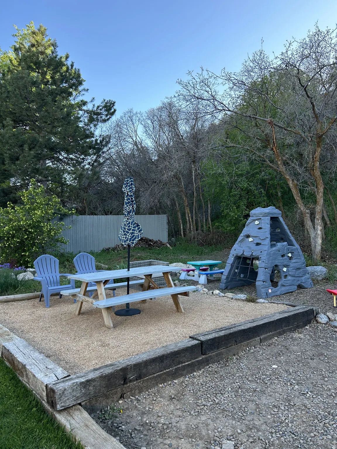 Outdoor patio featuring a gravel surface, wood timber retaining wall, and mature perimeter trees