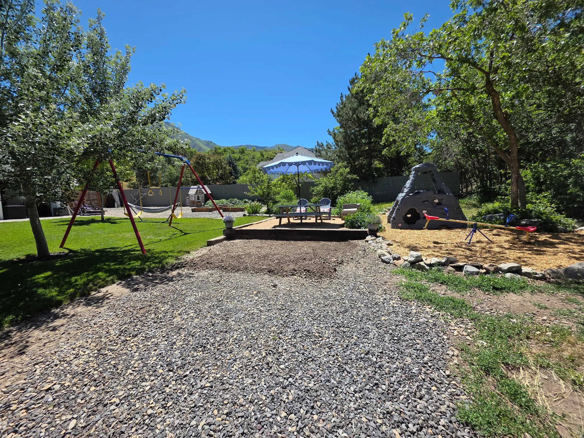 Expansive backyard featuring a green lawn, mature trees, a gravel pathway, and a wood chip play area