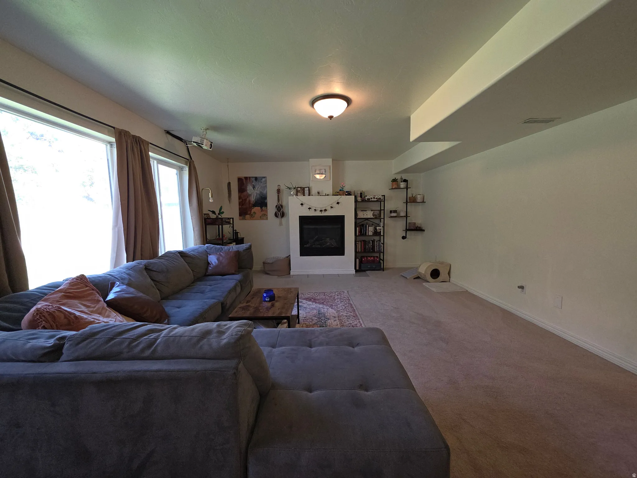 Spacious living area featuring a built-in fireplace with a white mantel, large windows, recessed ceiling beams, and light-toned carpet flooring
