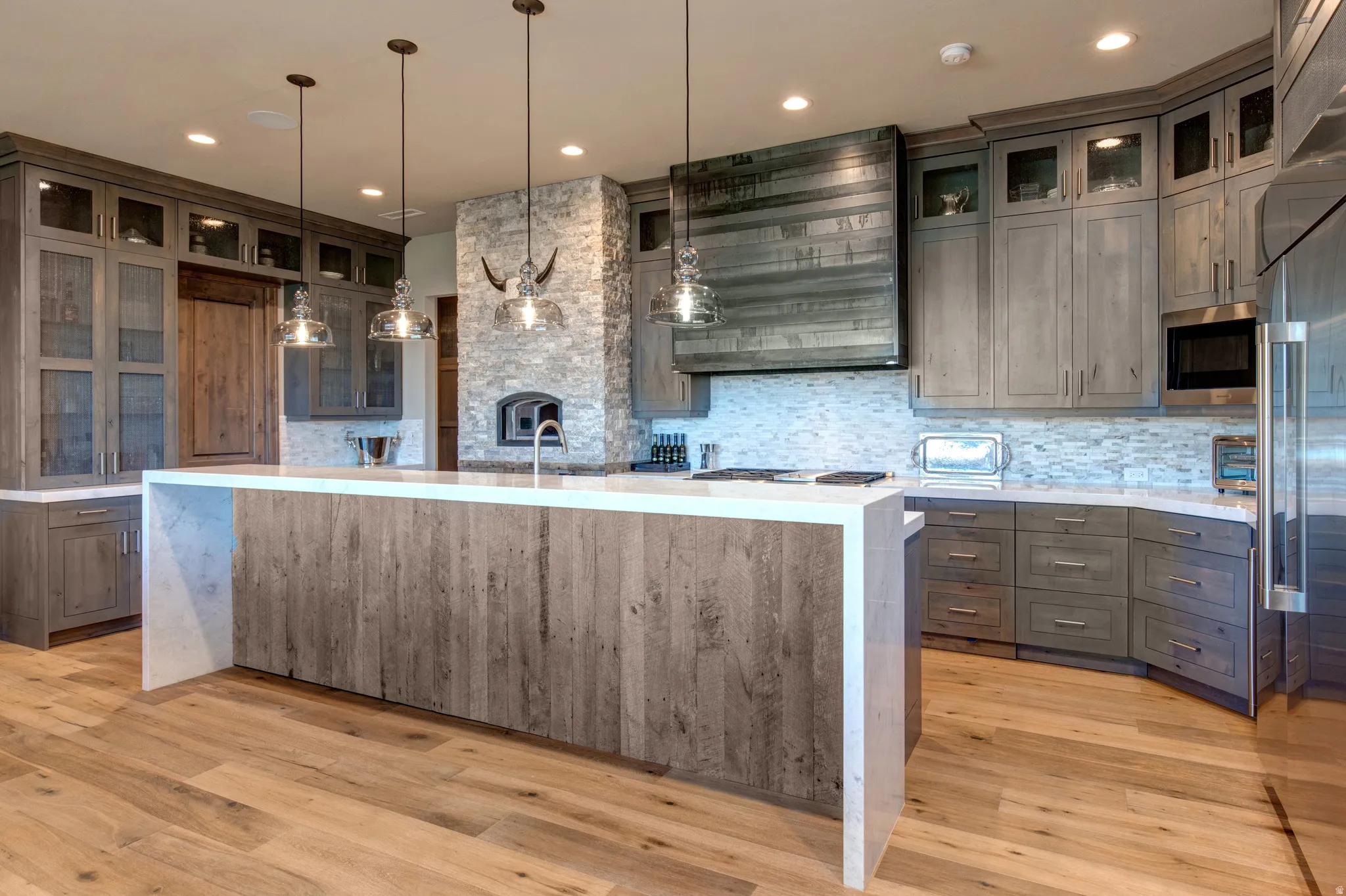 Spacious kitchen featuring a large island with wood-finish paneling, light wood-finish flooring, and light-toned cabinetry with glass-front uppers