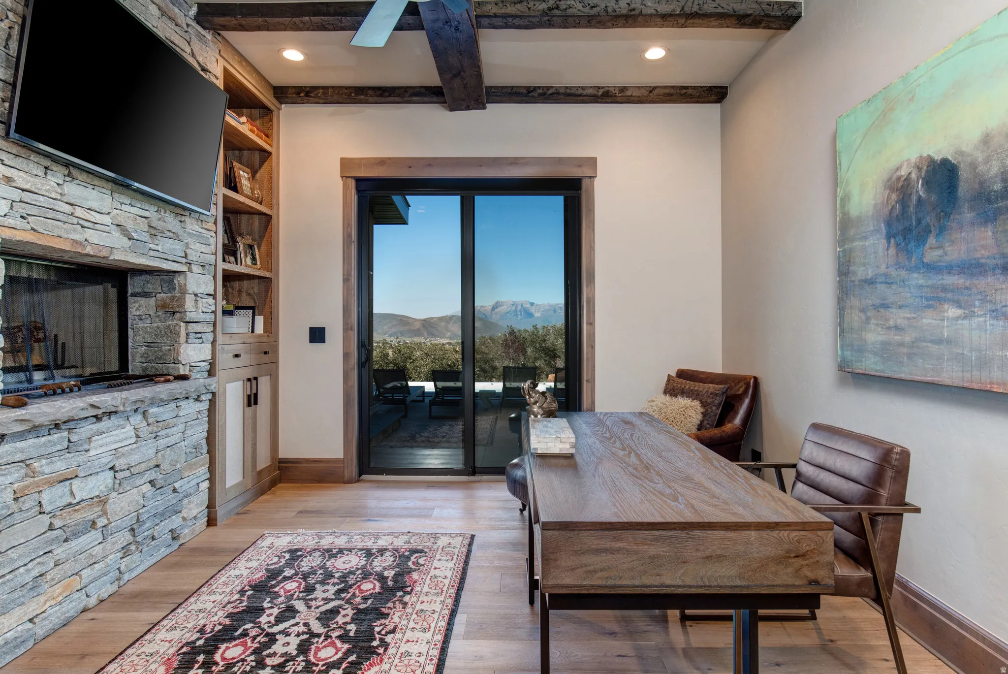 Stone-clad fireplace with a built-in entertainment center, wood-finish flooring, exposed ceiling beams, and a sliding glass door