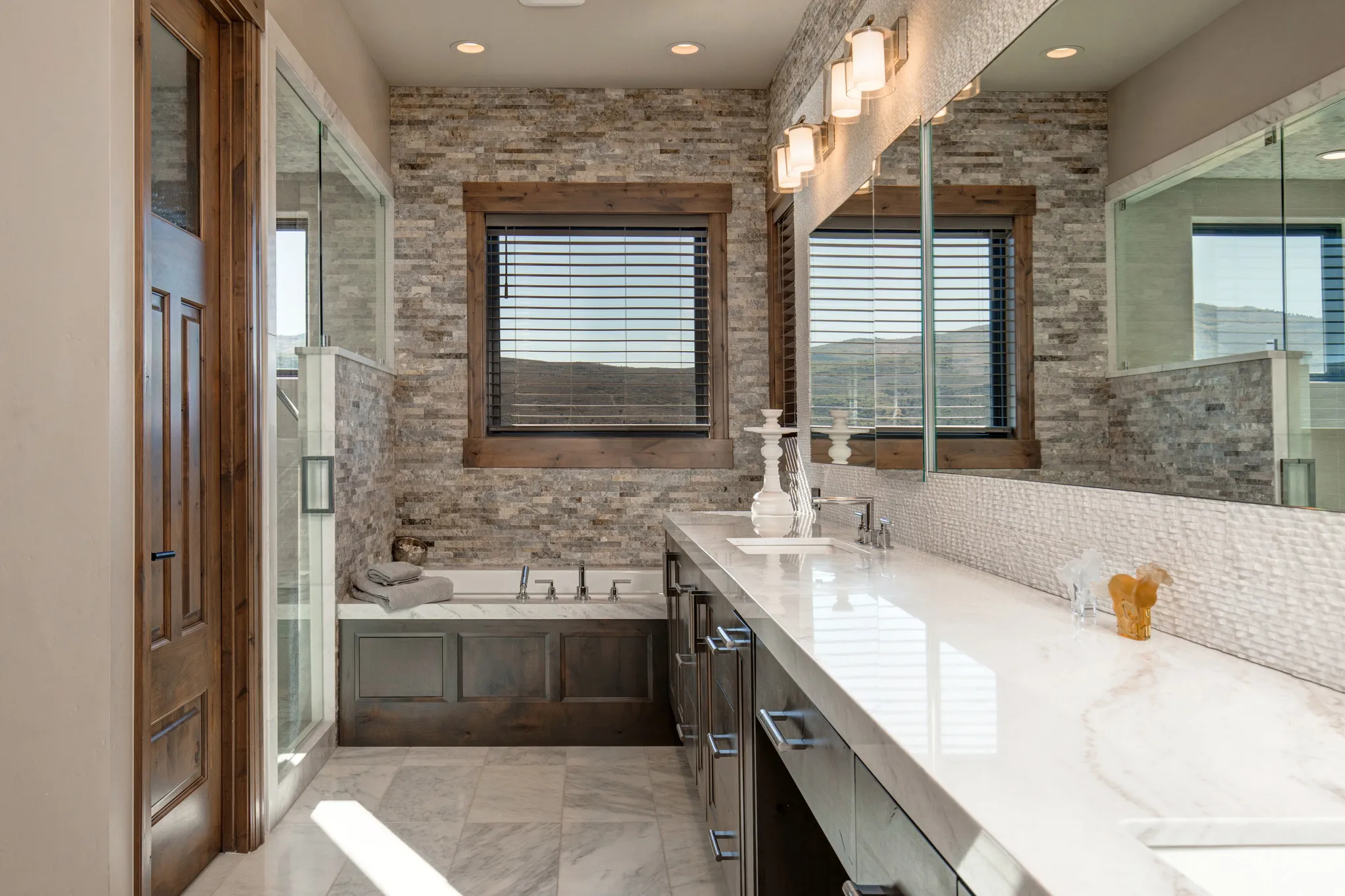 Spacious bathroom featuring a dual vanity with a light-toned countertop, integrated soaking tub, stone accent wall, glass-enclosed shower, and wood-paneled door
