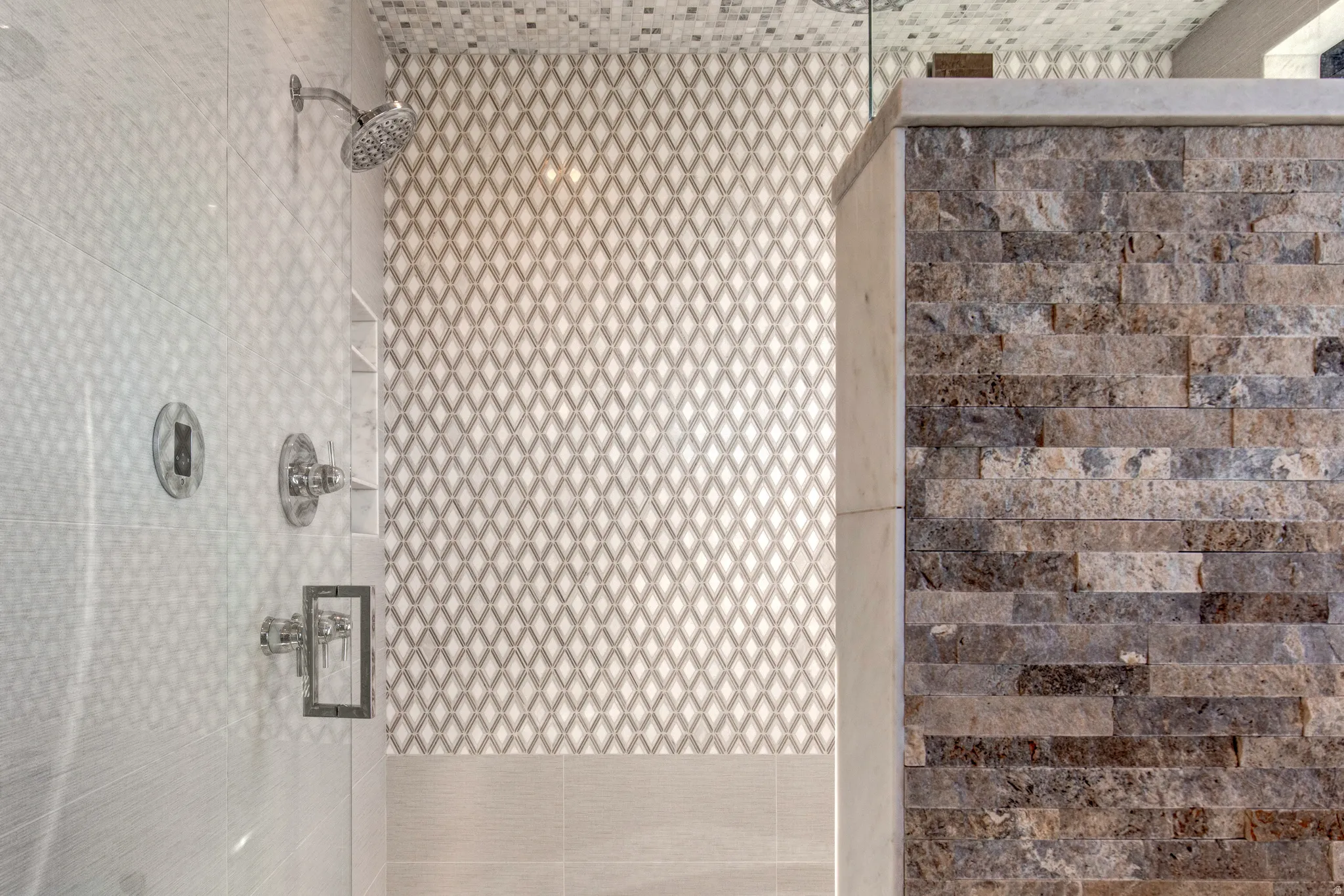 Shower with polished chrome fixtures, diamond pattern mosaic wall tile, built-in recessed shelving, and a stone-clad half wall