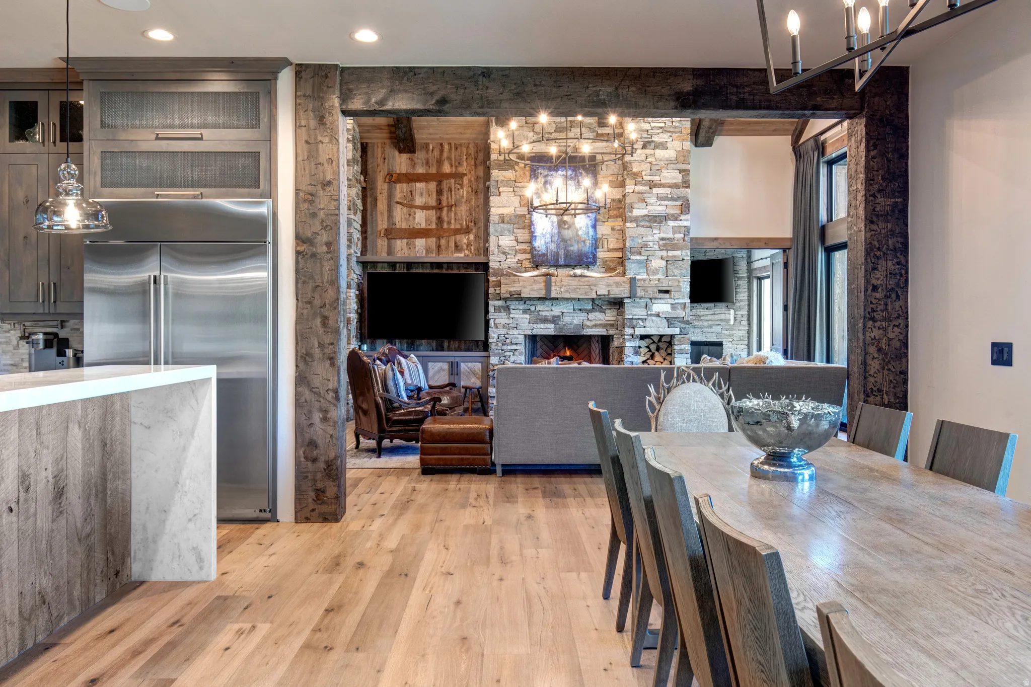 Open-concept design featuring wood-finish flooring, a stone-clad fireplace, exposed wood beams, and a substantial dining table