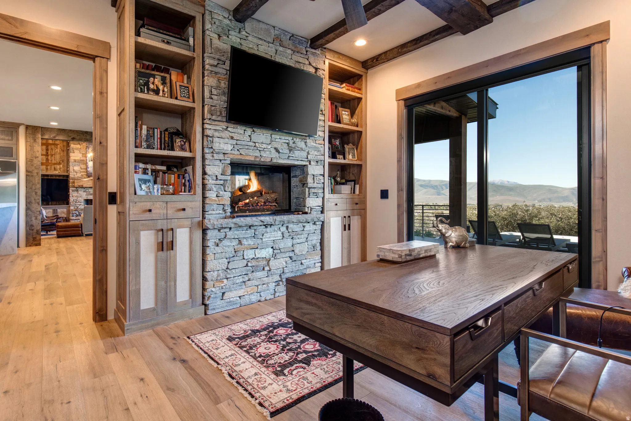 Spacious interior room featuring a stone fireplace, built-in shelving, wood-finish flooring, exposed ceiling beams, and sliding glass doors opening to outdoor scenery