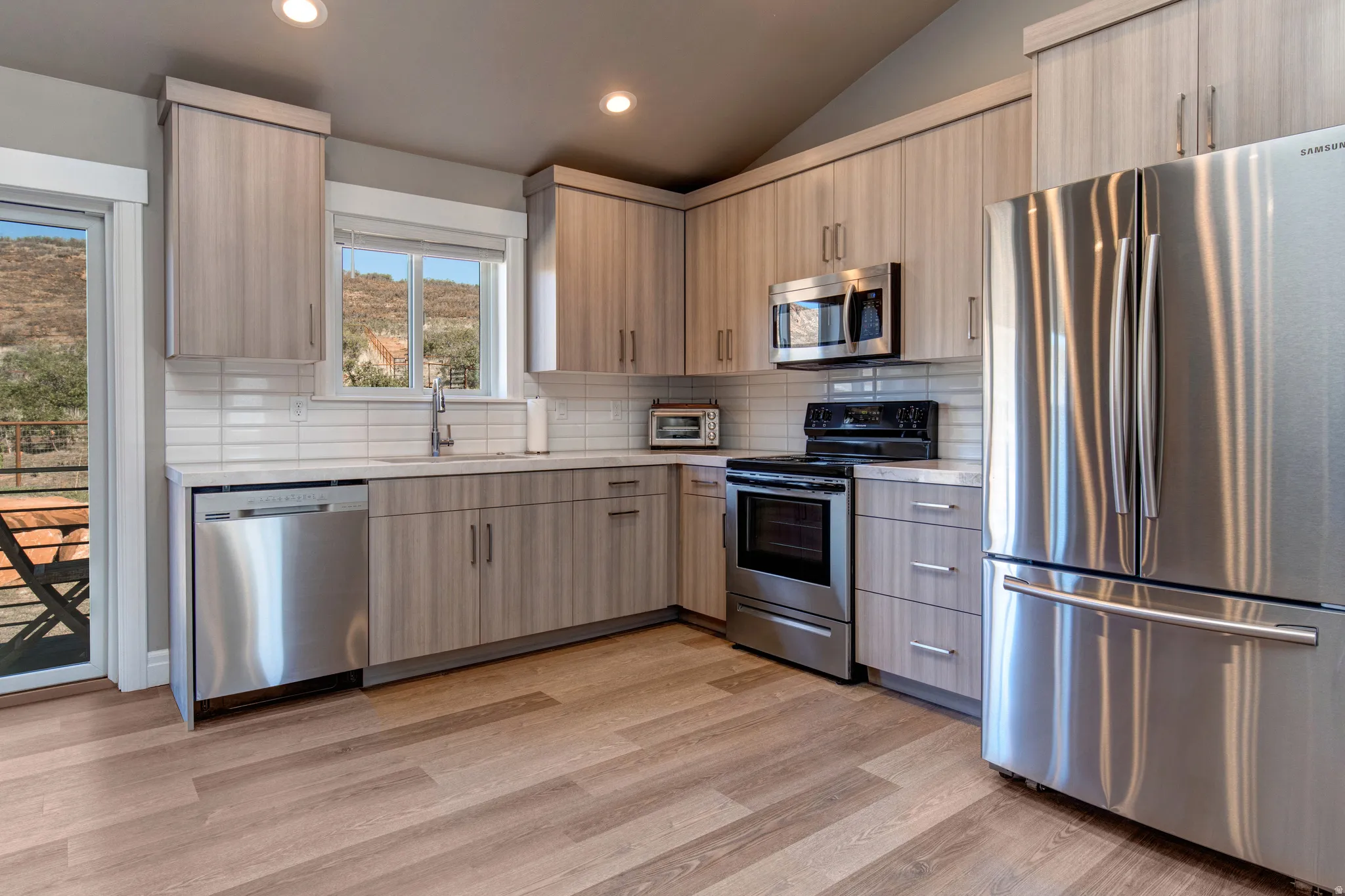 Modern kitchen featuring wood-finish cabinetry, stainless steel appliances, a white subway tile backsplash, light-toned countertops, and wood-finish flooring