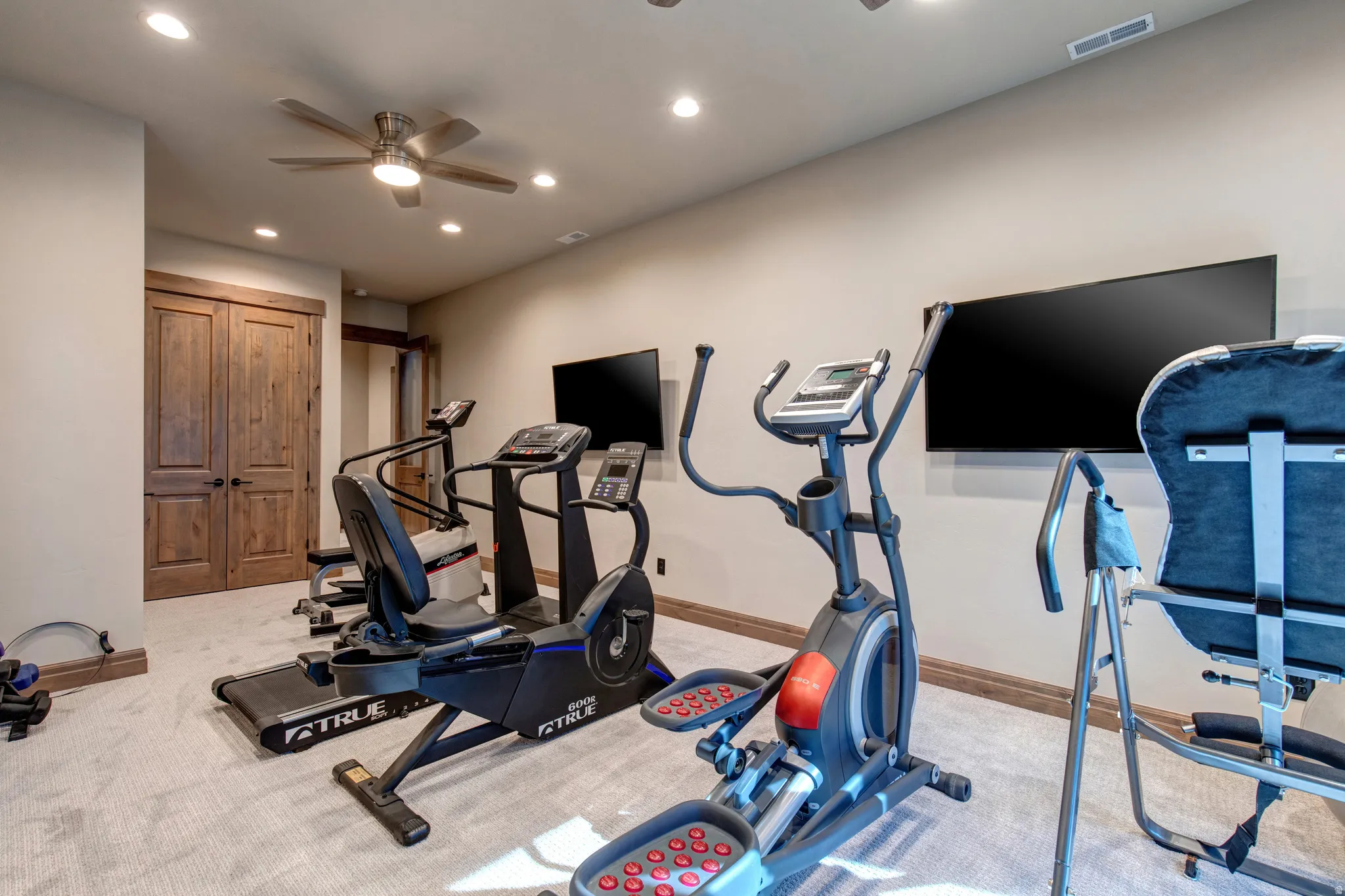 Dedicated exercise space featuring light-colored carpeting, recessed lighting, a ceiling fan, and two wall-mounted televisions