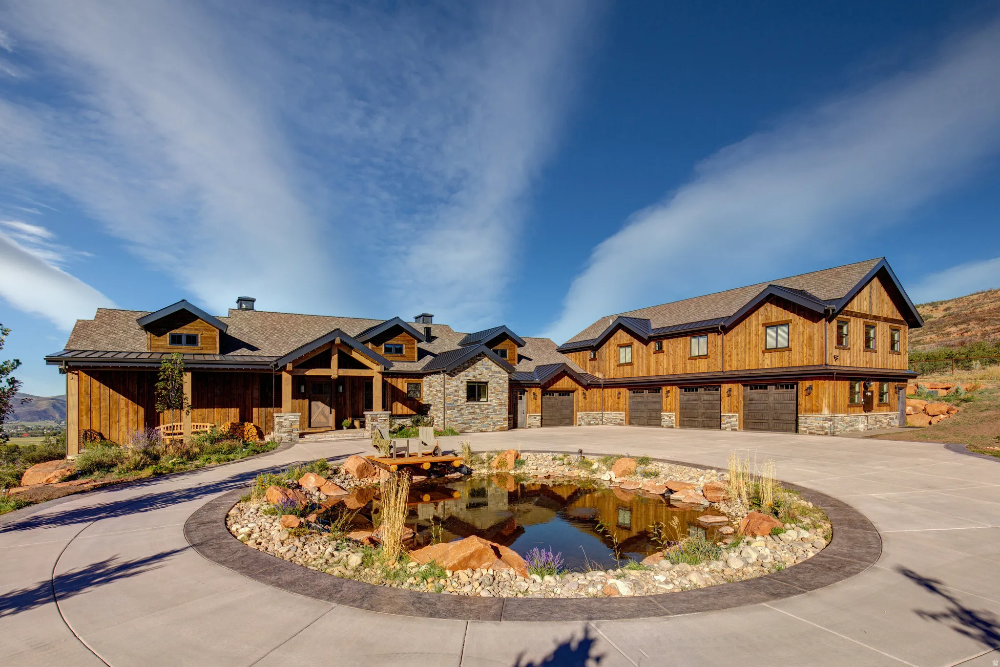 Grand estate featuring a circular driveway with a central water feature, a wood-sided exterior, stone accents, and multiple garage doors