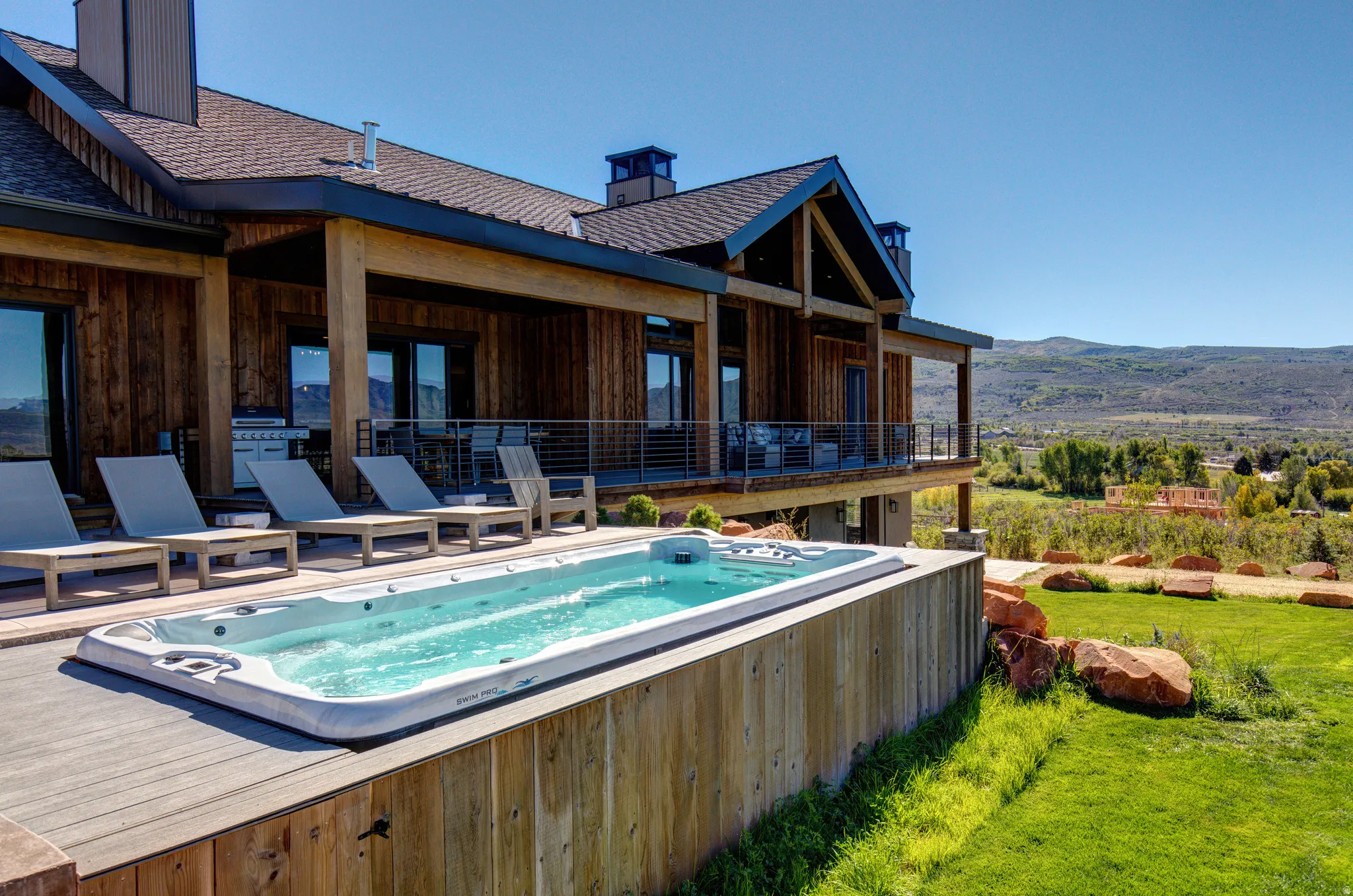 Expansive outdoor living area featuring a swim spa, wood-finish decking, and a covered patio with a built-in grill