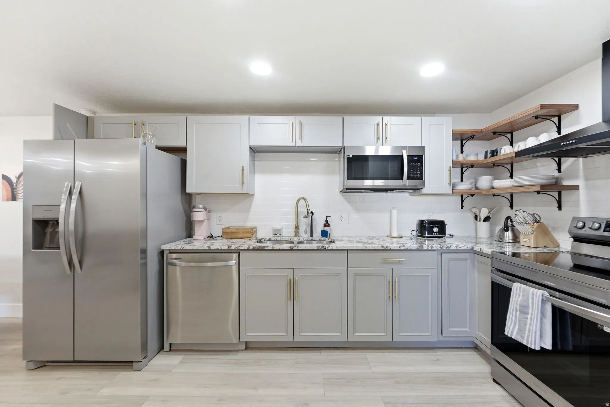Modern kitchen featuring light gray cabinetry with gold-tone hardware, stainless steel appliances, white subway tile backsplash, and light-toned wood-finish flooring