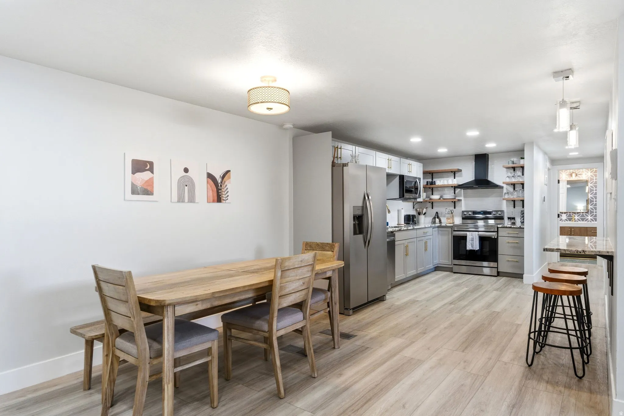 Open-concept kitchen and dining area featuring light wood-finish flooring, stainless steel appliances, light gray cabinetry, and a granite-finish breakfast bar