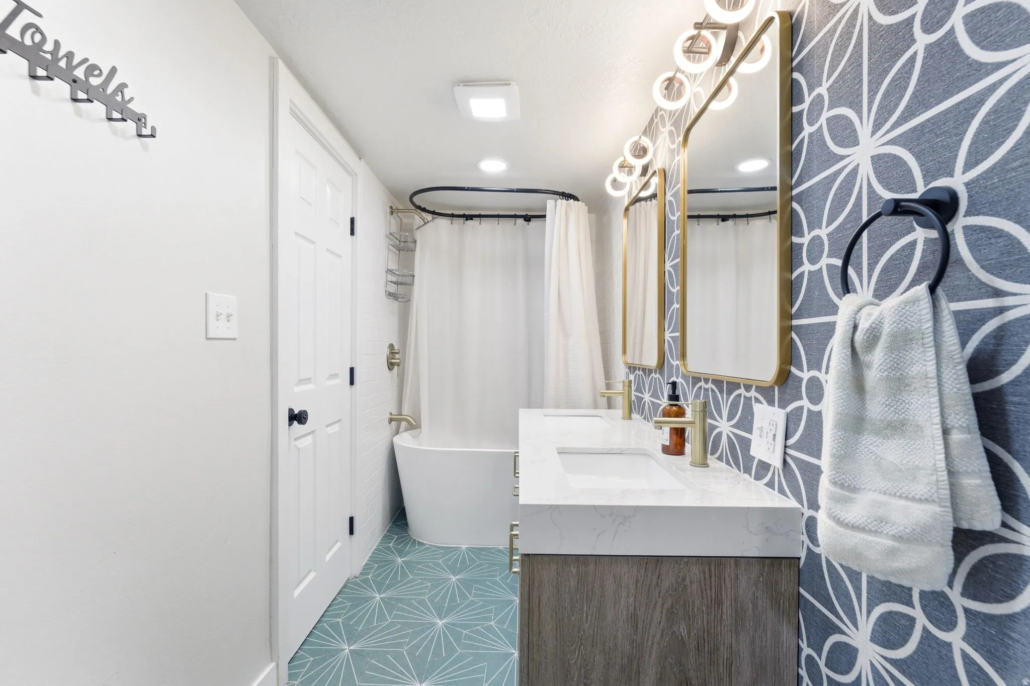 Bathroom featuring a dual vanity with integrated sinks, gold-framed mirrors, and patterned wallcovering
