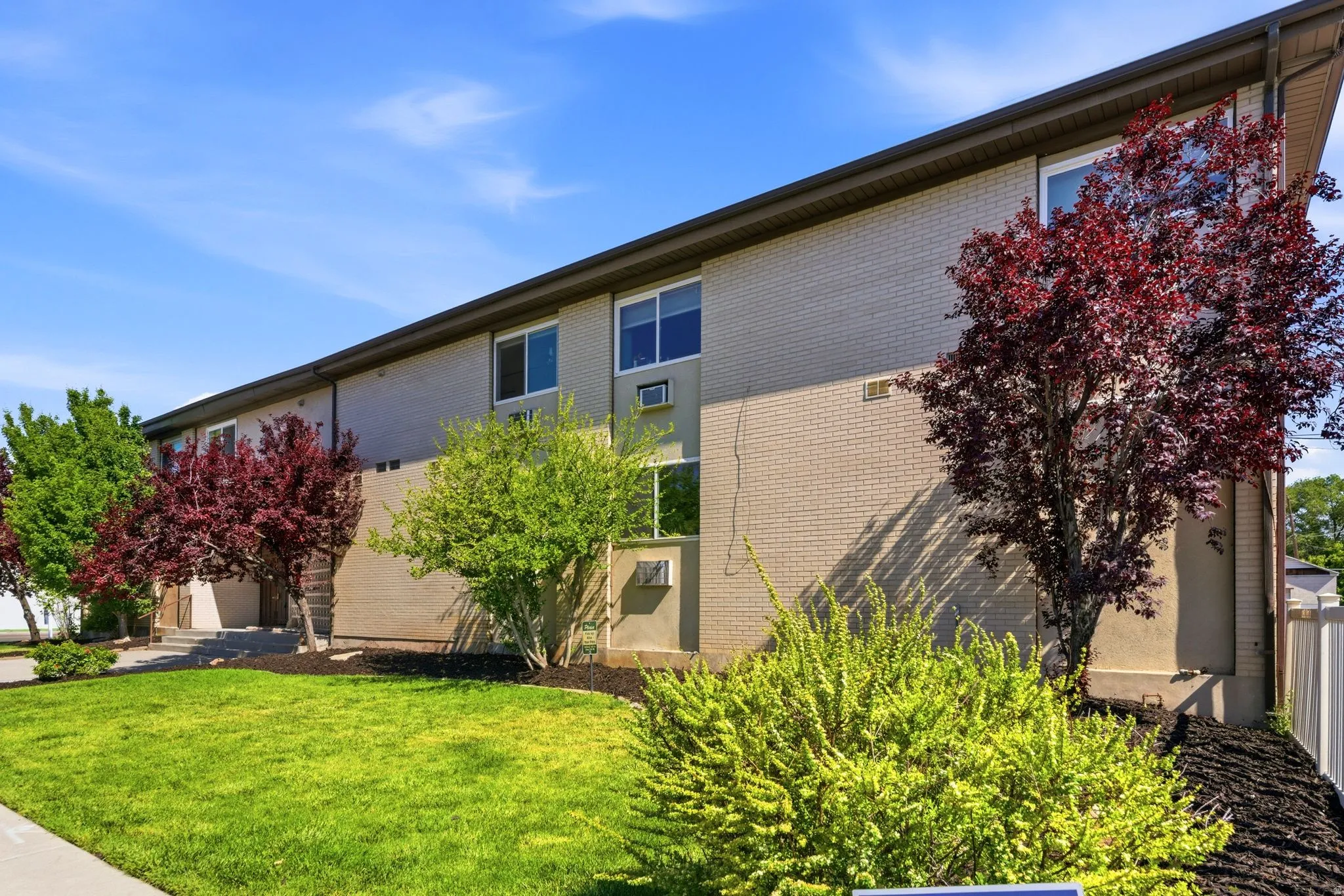 Light brick and stucco multi-story building featuring multiple windows, a sloped roofline, and established landscaping