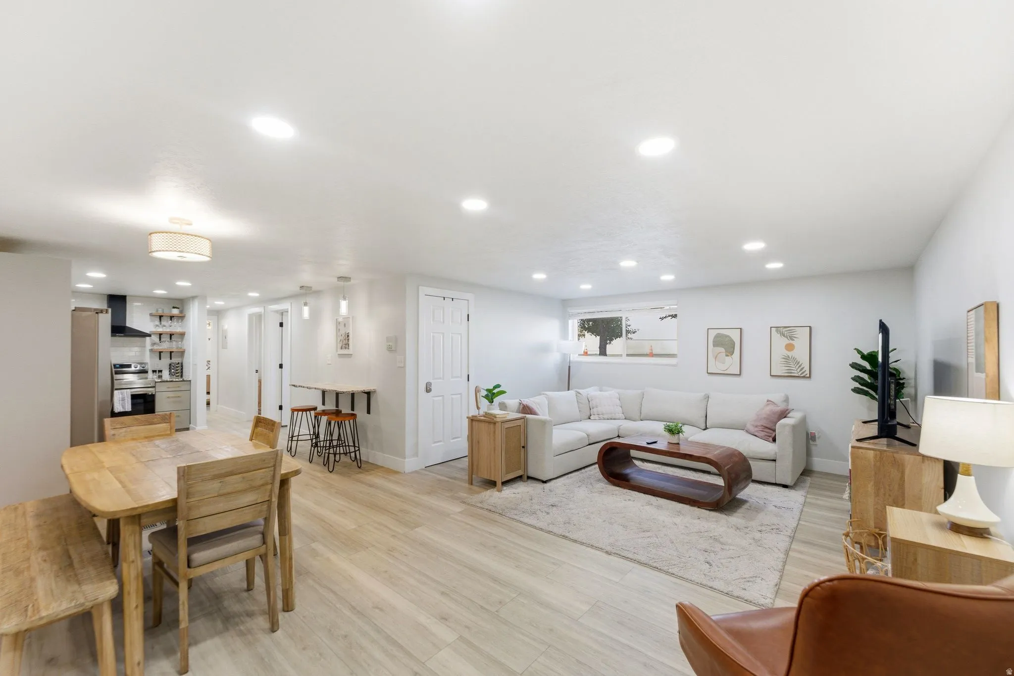 Open-concept living space featuring light wood-finish flooring, recessed lighting, a built-in kitchen area with a black range hood, and a dining area with a built-in wall-mounted table