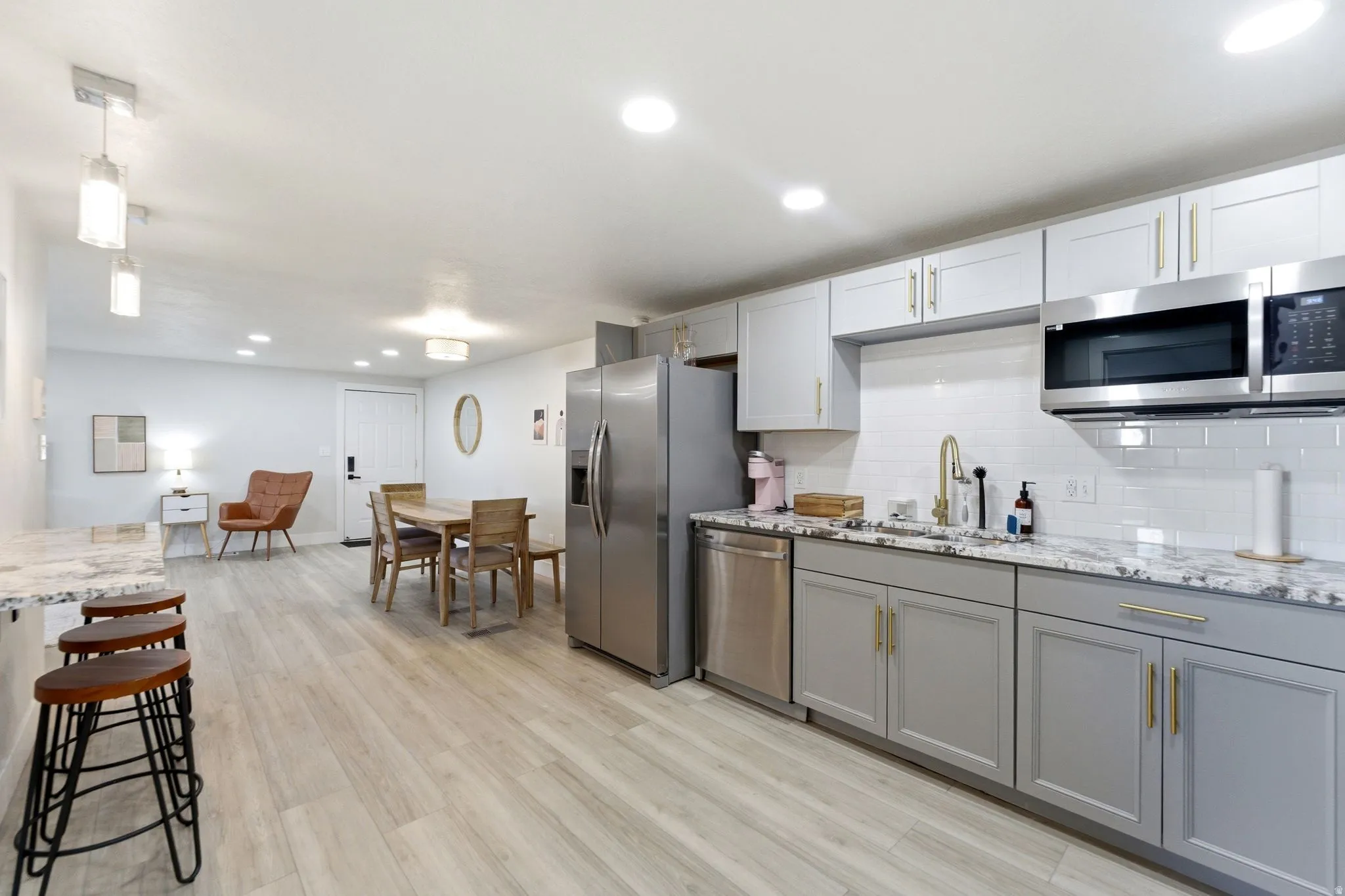 Open-concept kitchen featuring stainless steel appliances, light wood-finish flooring, two-tone cabinetry with gold-tone hardware, white subway tile backsplash, and stone countertops