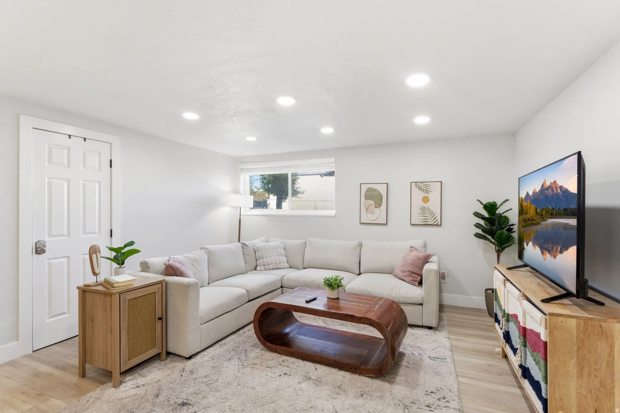 Spacious living area featuring light wood-finish flooring, recessed lighting, a window providing natural light, a white paneled door, and neutral wall tones