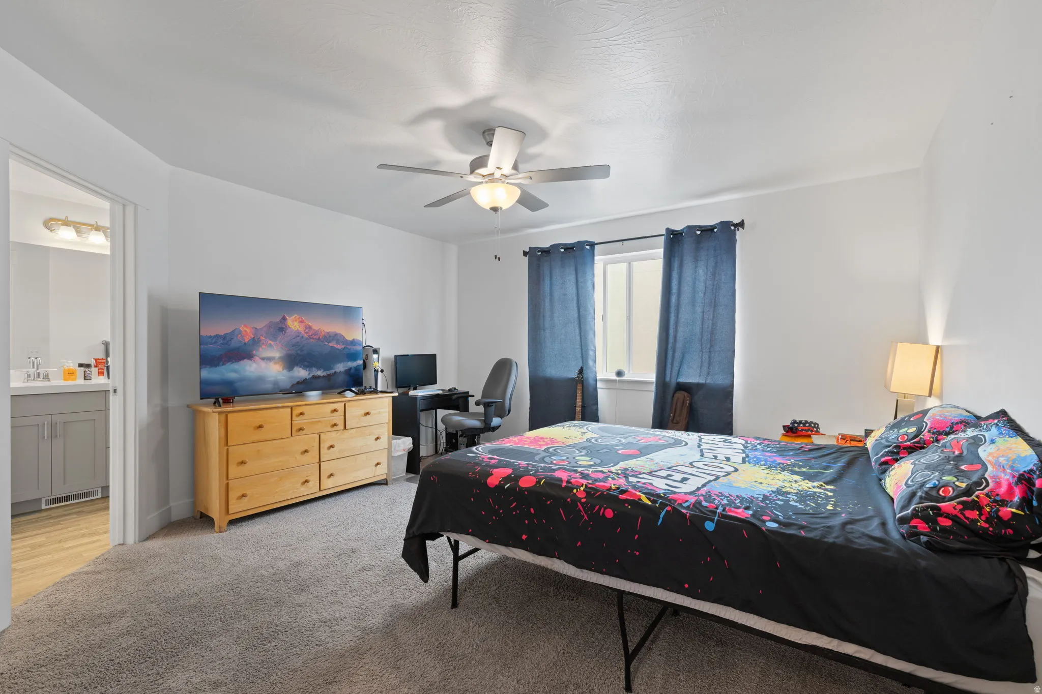 Carpeted bedroom with a ceiling fan light fixture, a window with curtains, and an entry to an adjacent bathroom featuring a vanity with a sink and wall-mounted light fixture