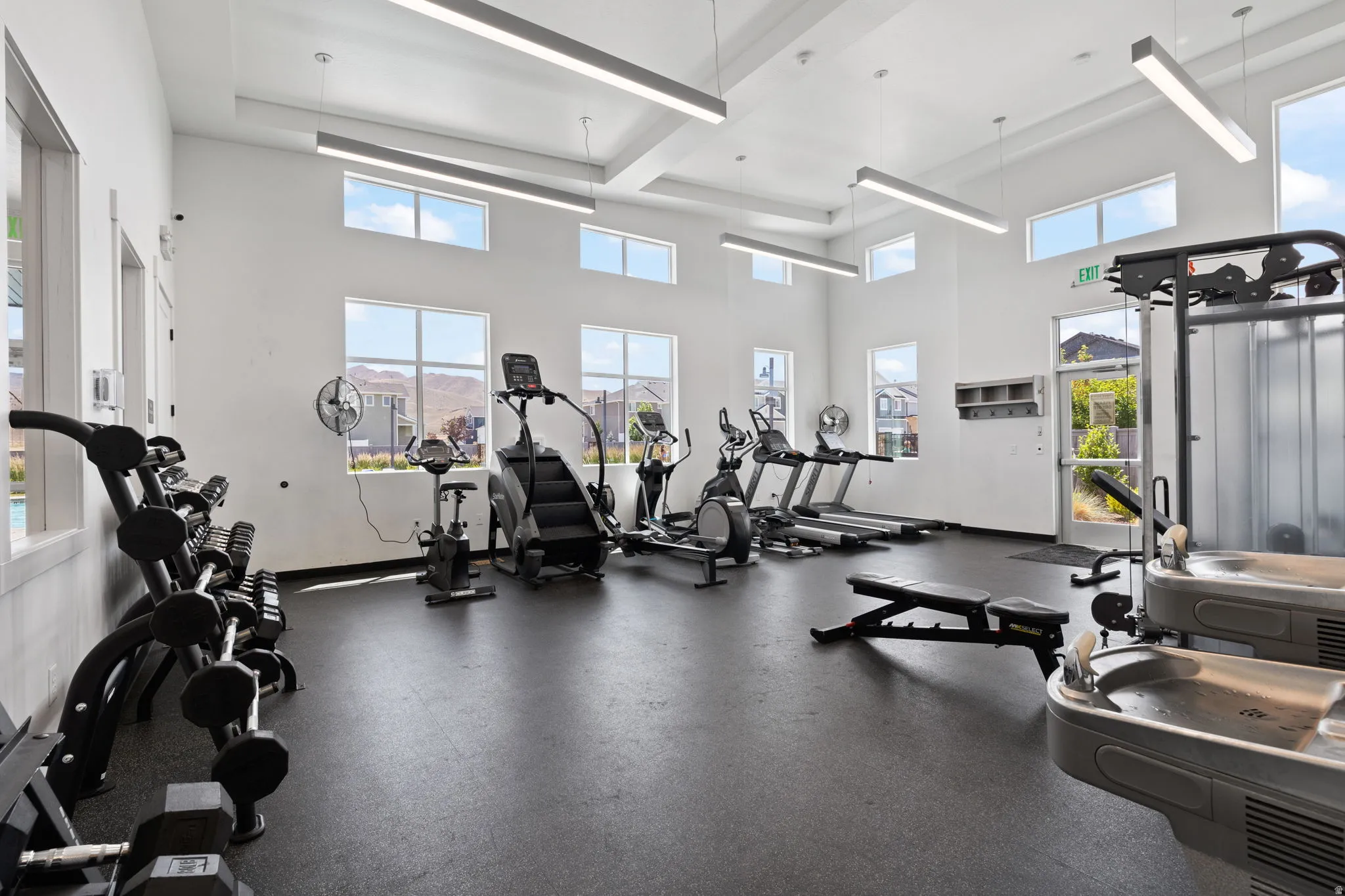 Fitness center featuring high ceilings, ample natural light from multiple windows, durable rubber flooring, and modern linear pendant lighting
