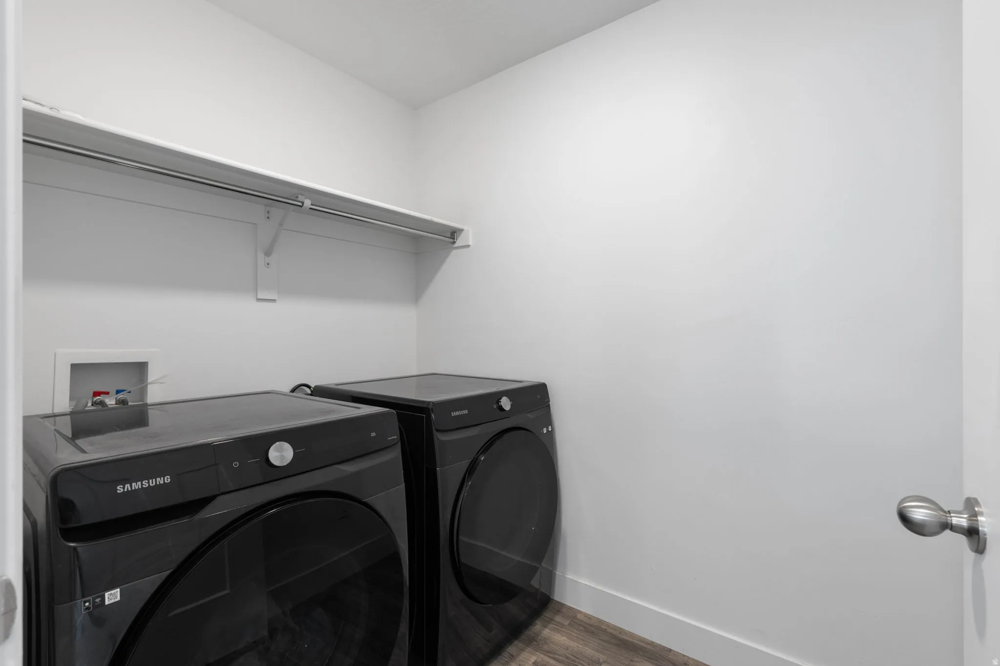 Dedicated laundry area featuring modern black-finish appliances, wood-finish flooring, white walls, a white baseboard, and a white storage shelf with a hanging rod