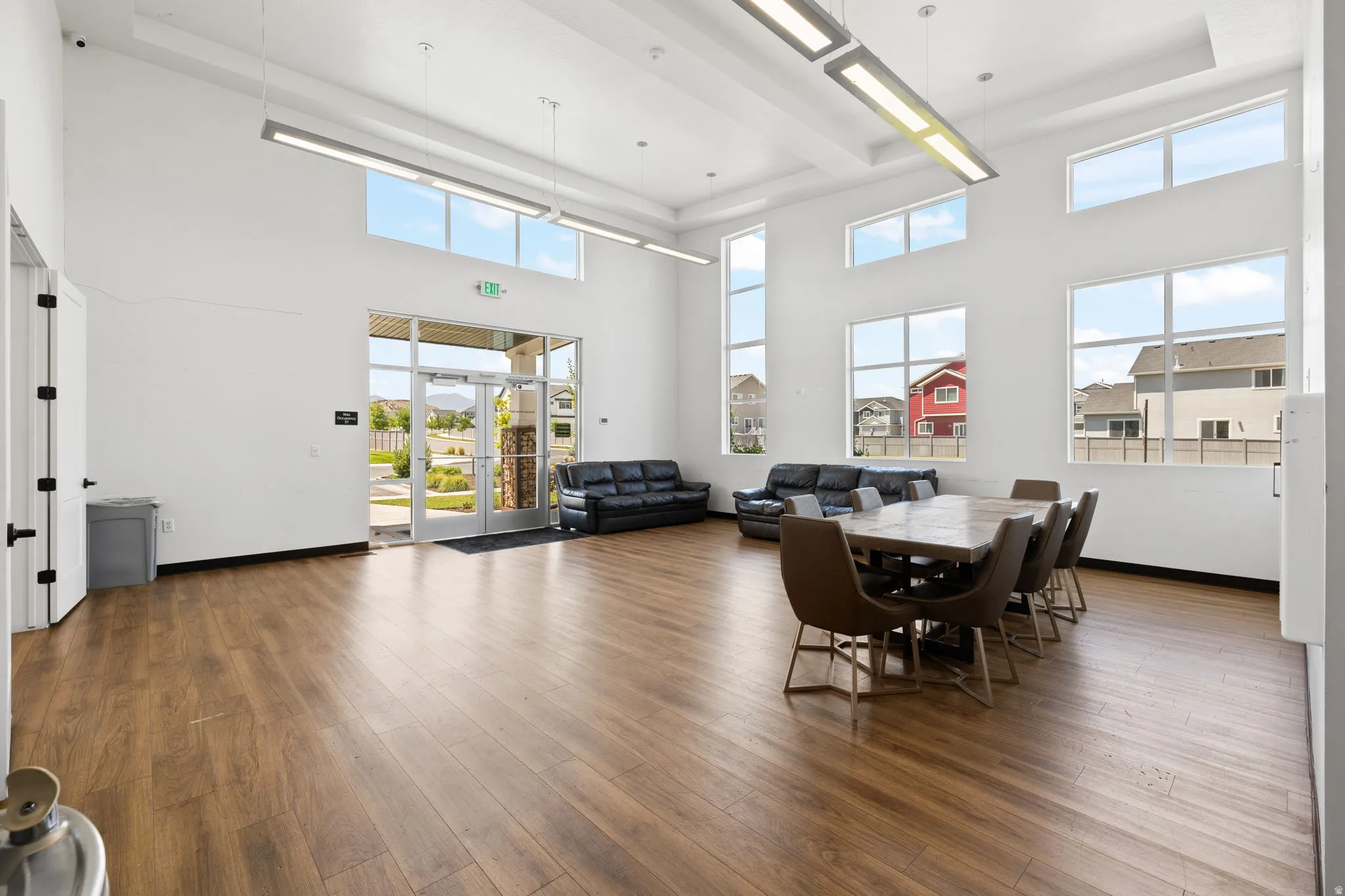 Spacious common area featuring high ceilings, wood-finish flooring, abundant natural light from multiple large windows, modern overhead lighting, and an expansive entry with glass double doors