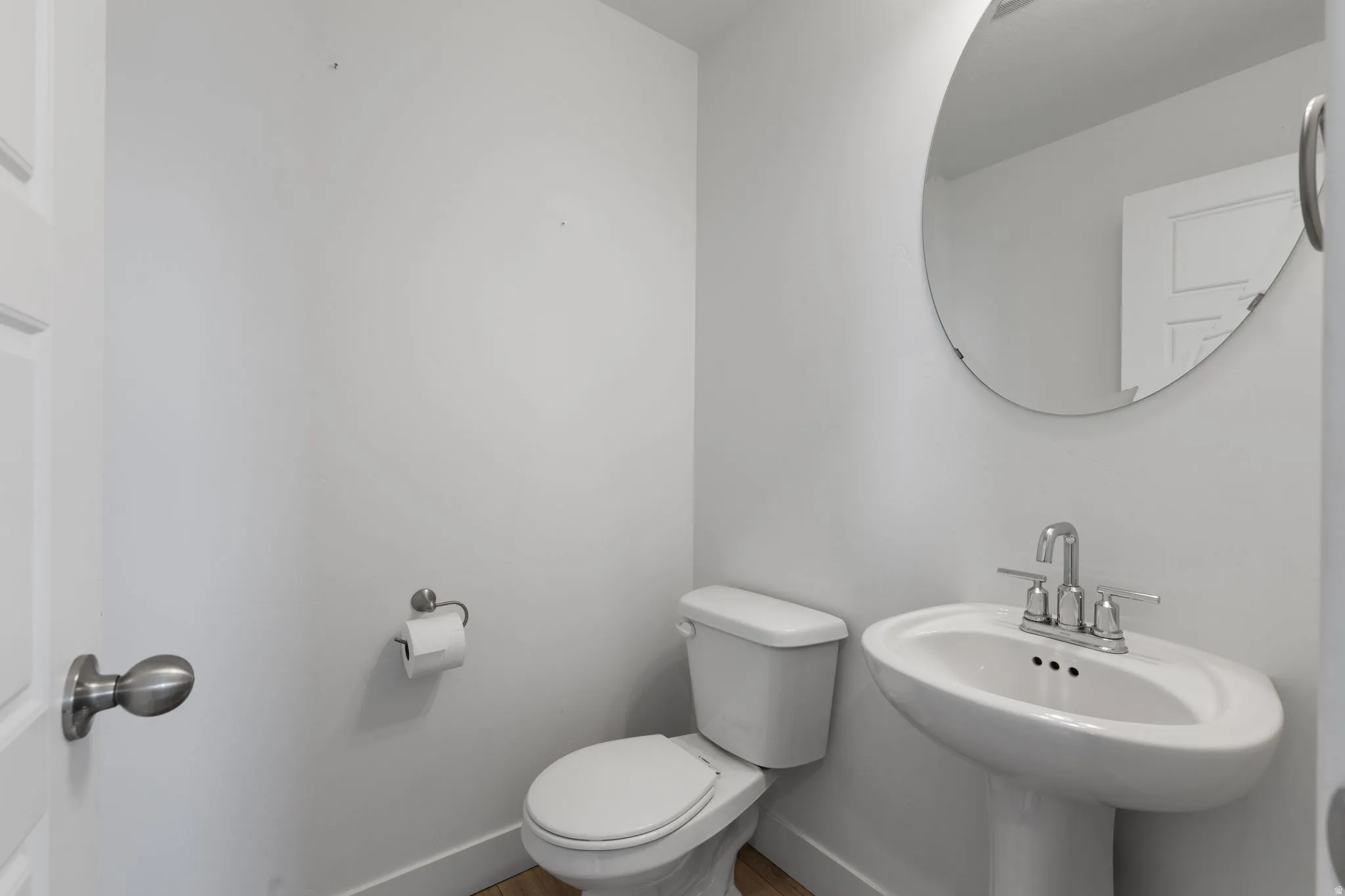 Pedestal sink with chrome faucet, circular mirror, and wood-finish flooring