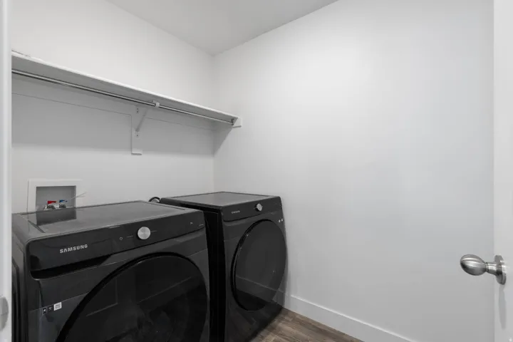 Dedicated laundry area featuring modern black-finish appliances, wood-finish flooring, white walls, a white baseboard, and a white storage shelf with a hanging rod