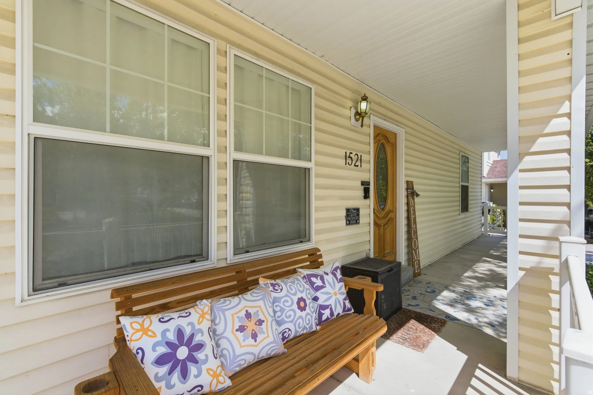 Covered front porch featuring cream-colored siding, a wooden front door with an oval glass insert, and a gold-finish wall lantern