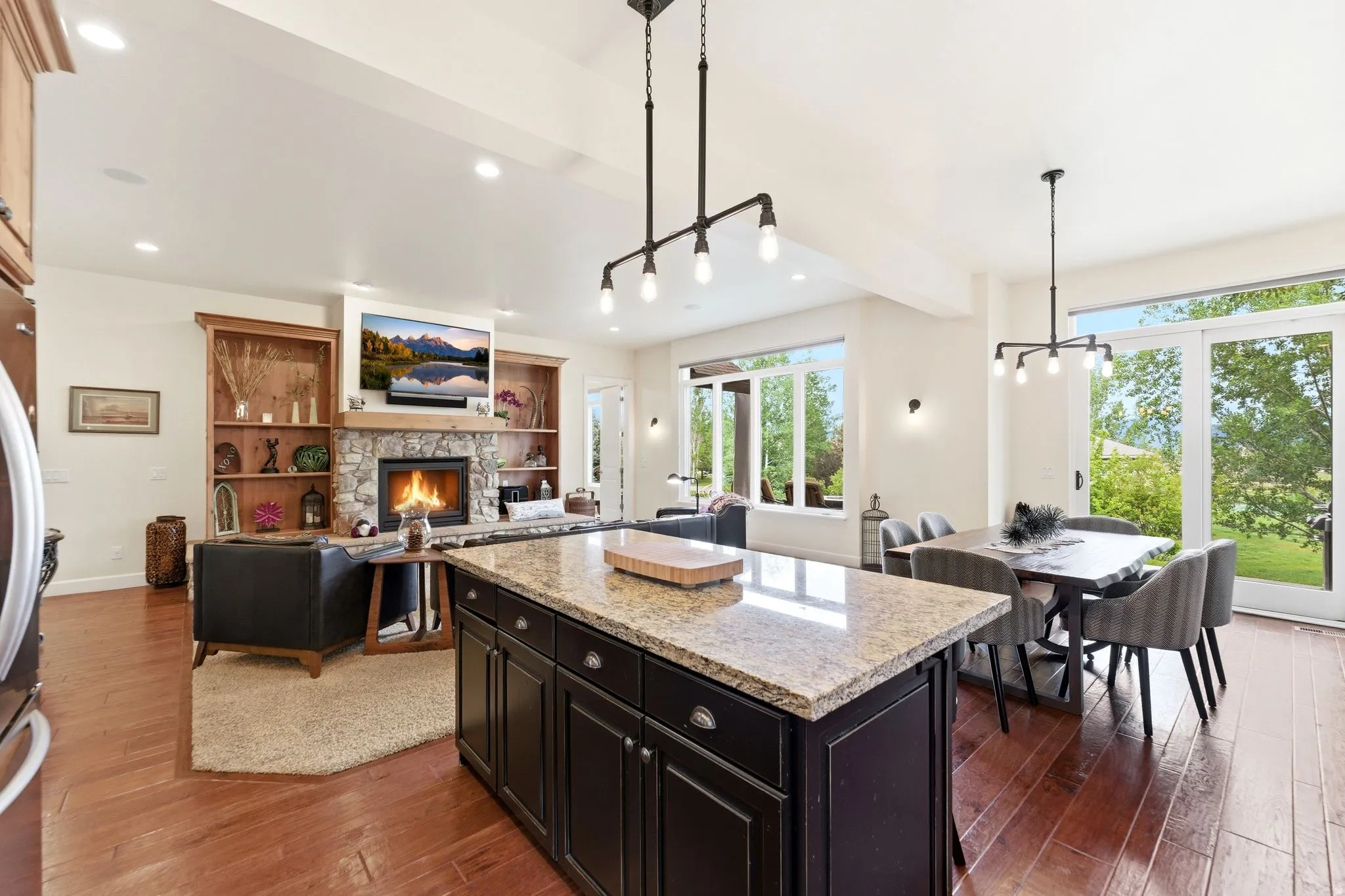 Open-concept living space featuring a central kitchen island with granite countertop, a stone fireplace with a wood mantle and built-in shelving, wood-finish flooring, a dining area with pendant lighting, and large glass sliding doors providing exterior a