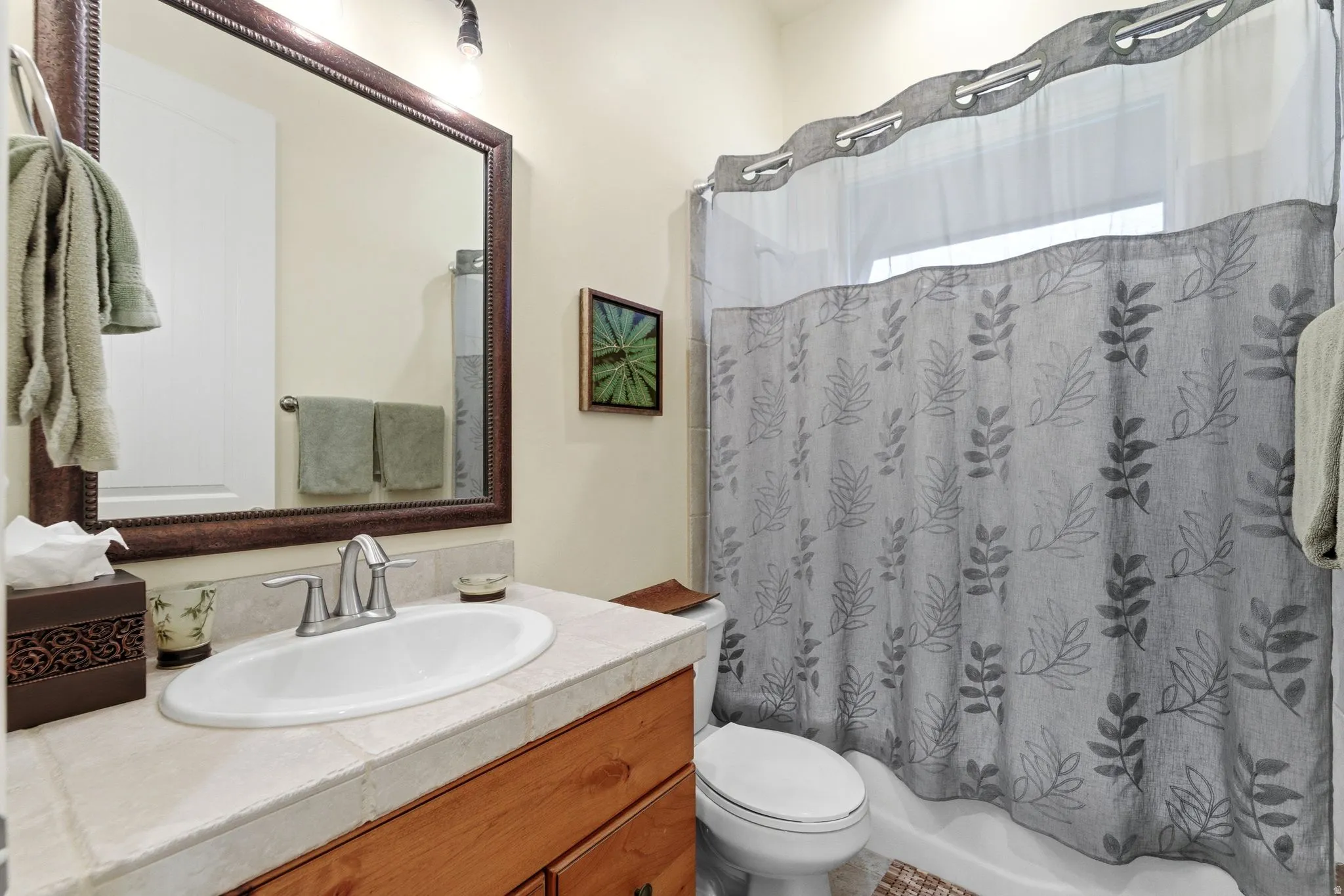 Bathroom featuring a white oval sink, tiled countertop, wood-finish vanity, and a shower-over-tub with a patterned curtain