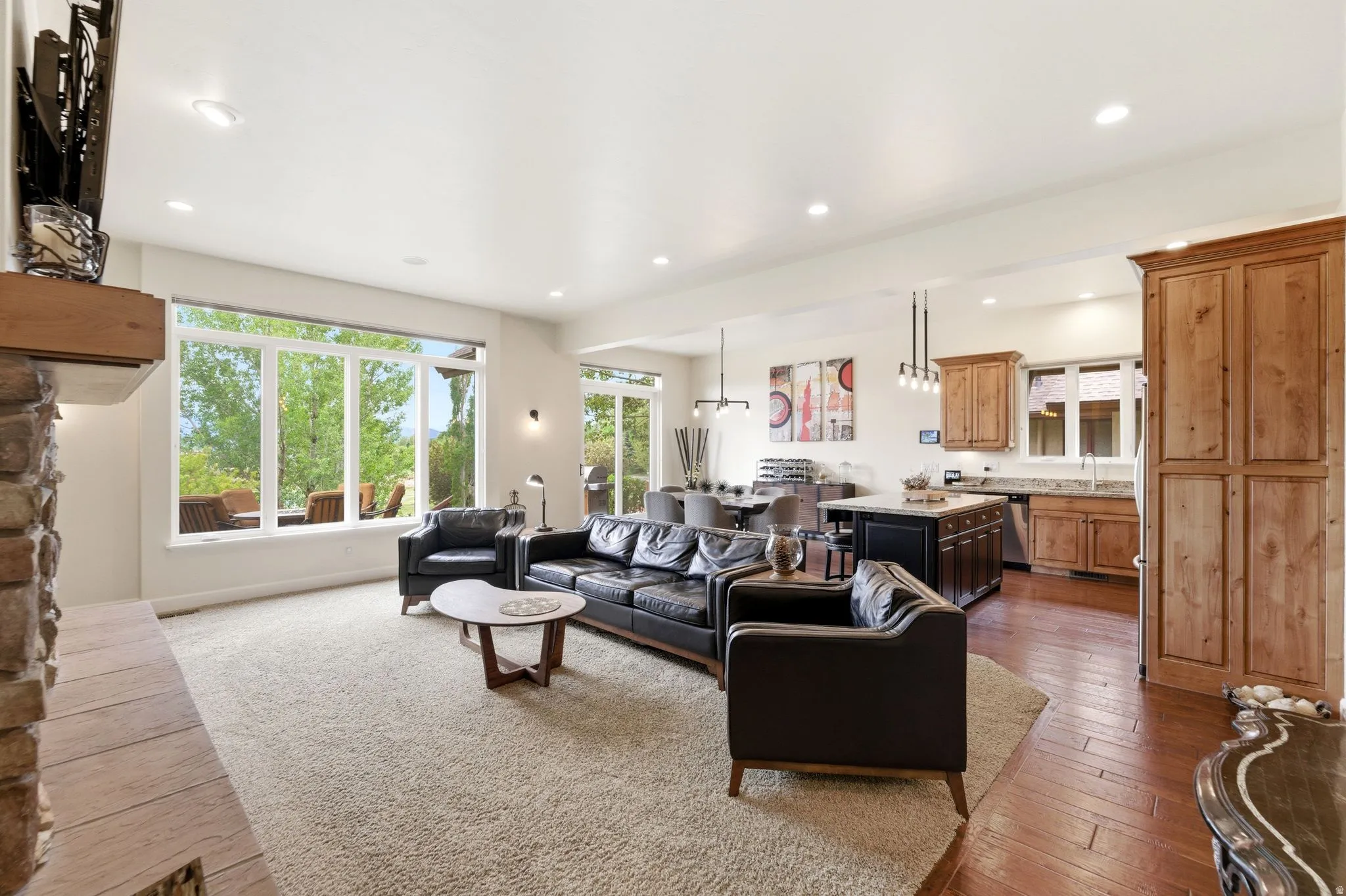 Open-concept living space featuring a stone fireplace with a wood mantle, a wall of windows, recessed lighting, and wood-finish flooring in the kitchen and dining areas